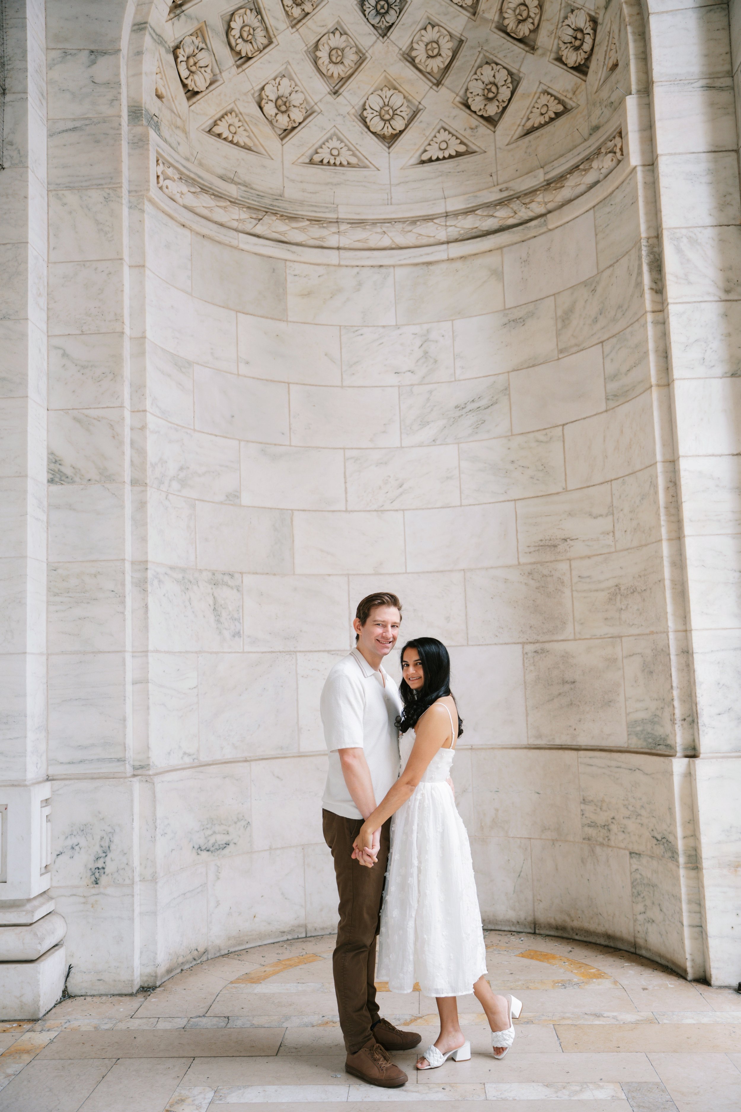 A smiling man and woman holding hands and standing close together, inside a large marble room with an ornate circular ceiling and matching wall pattern.