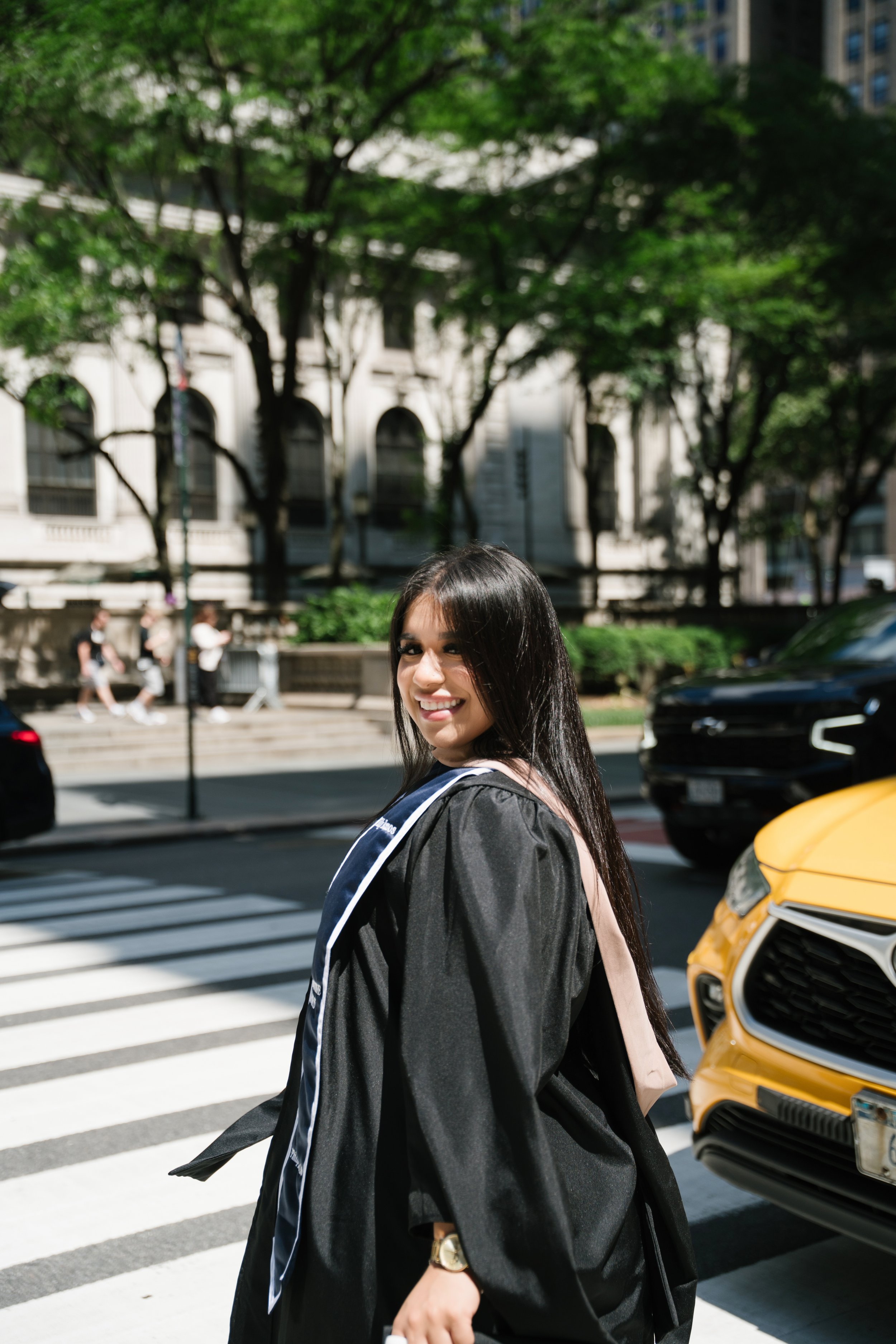 A young woman with long dark hair, wearing a graduation gown and carrying a backpack, walking across a street with a crosswalk, smiling at the camera. In the background, there are trees, a building, pedestrians, and cars.