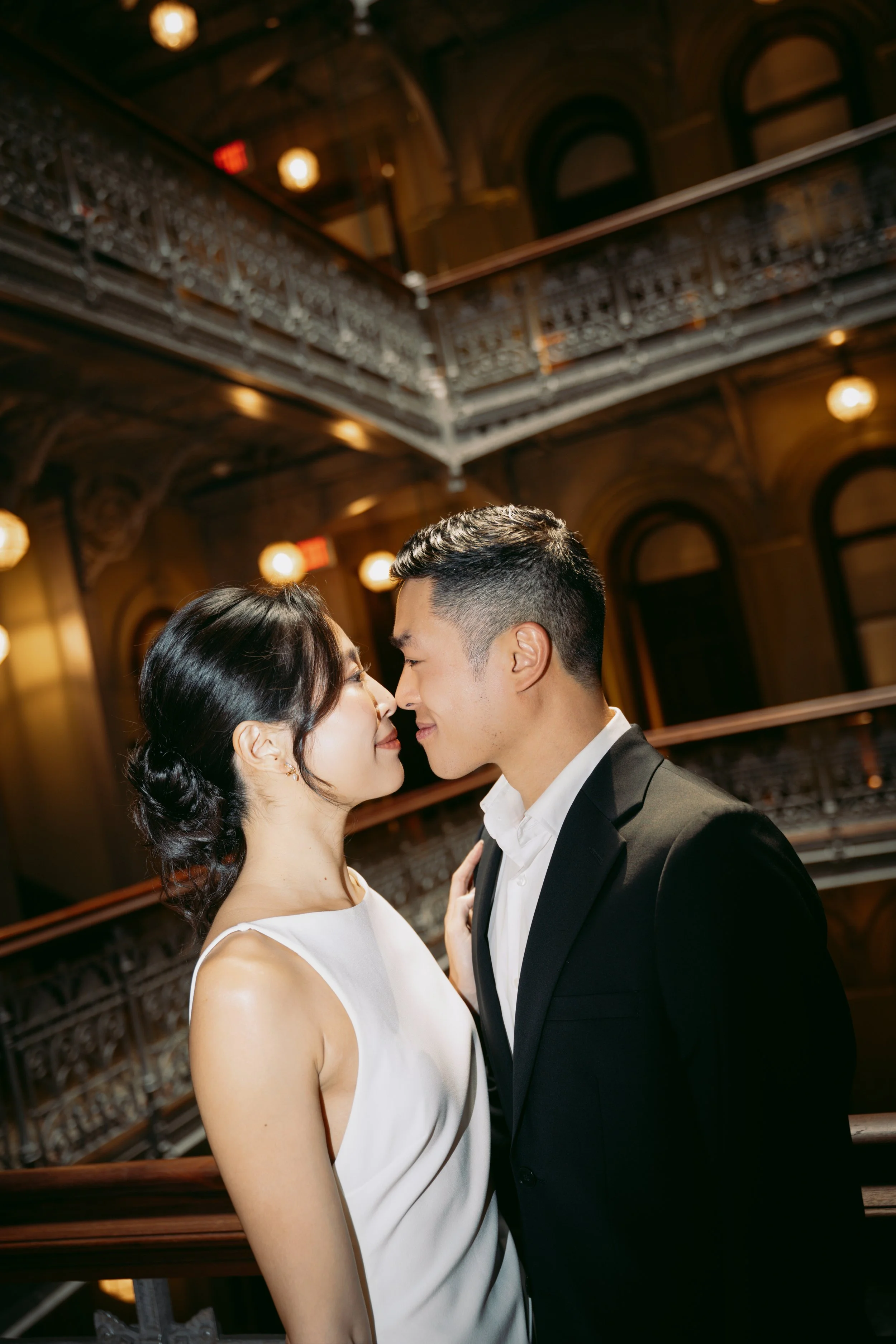 A couple, dressed in formal attire, is standing close together in an elegant, dimly lit venue with ornate architecture, possibly a wedding or special event.