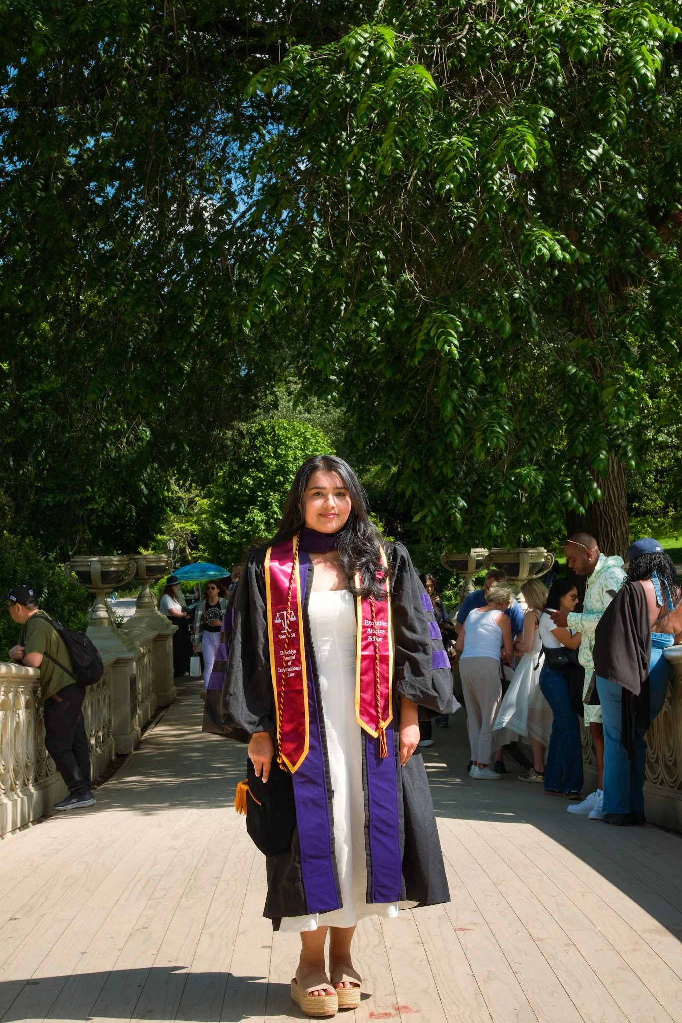 A woman dressed in a graduation gown and stole, standing outdoors with a crowd of people behind her, under a large leafy tree.
