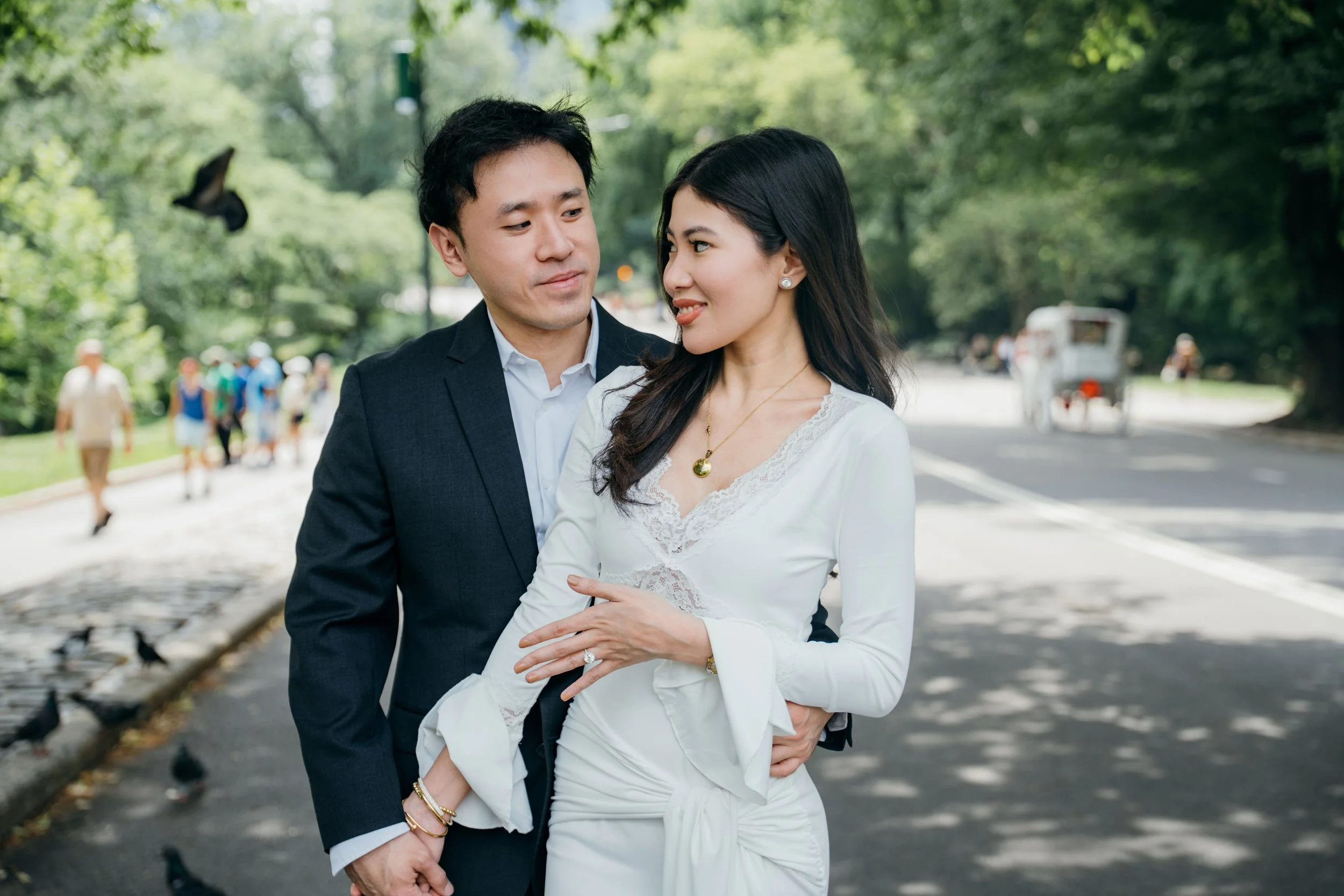 A couple, dressed formally, sharing an intimate moment outdoors on a tree-lined street with pedestrians and a horse-drawn carriage in the background.