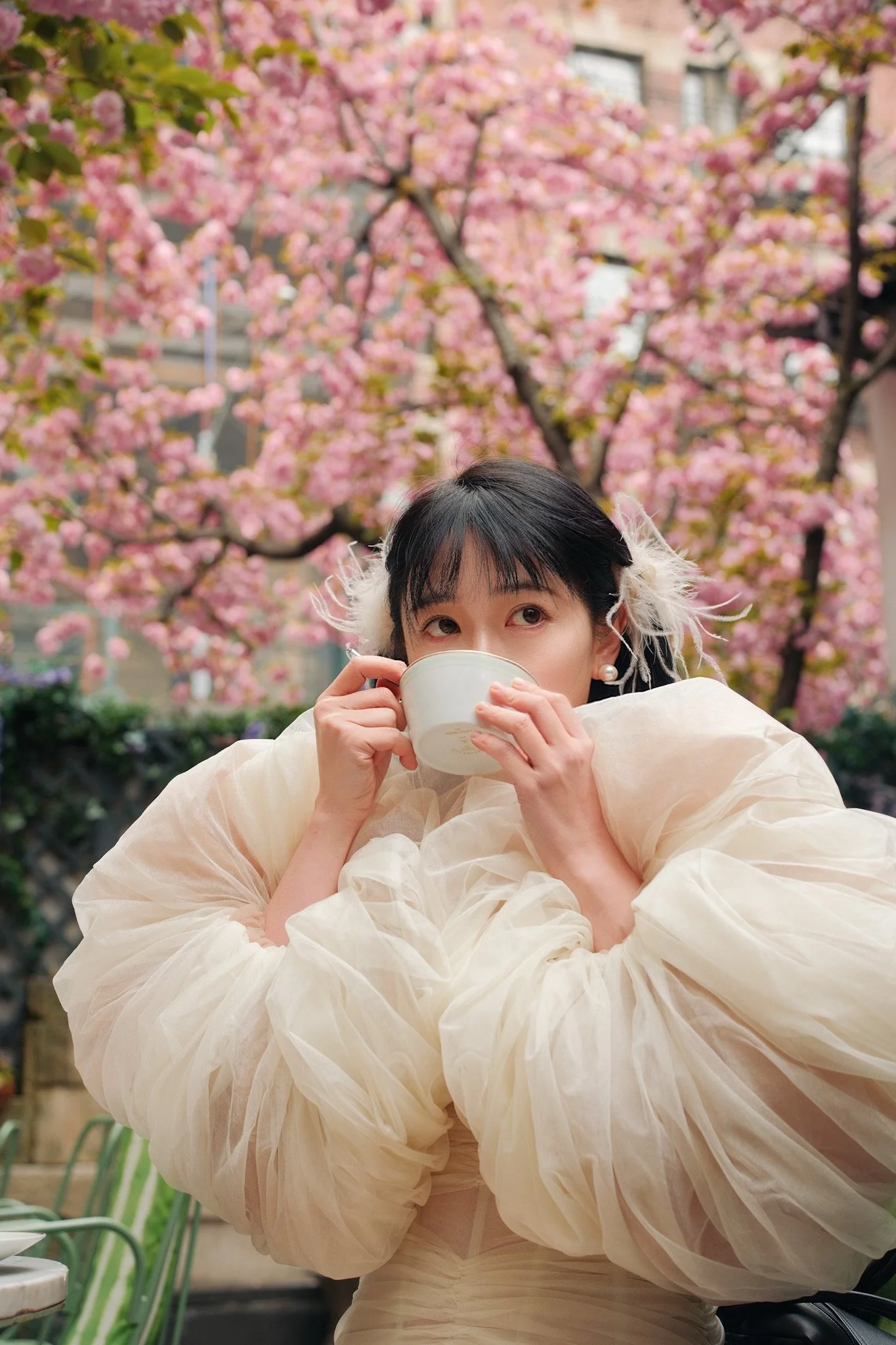 Woman with black hair and pearl earrings drinking from a white cup, wearing a white, ruffled, semi-transparent dress, standing outdoors with pink cherry blossoms in the background.