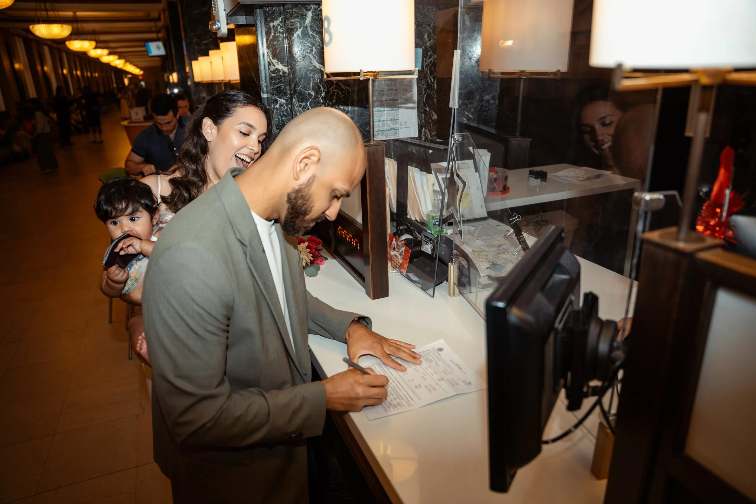 A man signing a document at a front desk in a hotel lobby with two women and a child nearby.