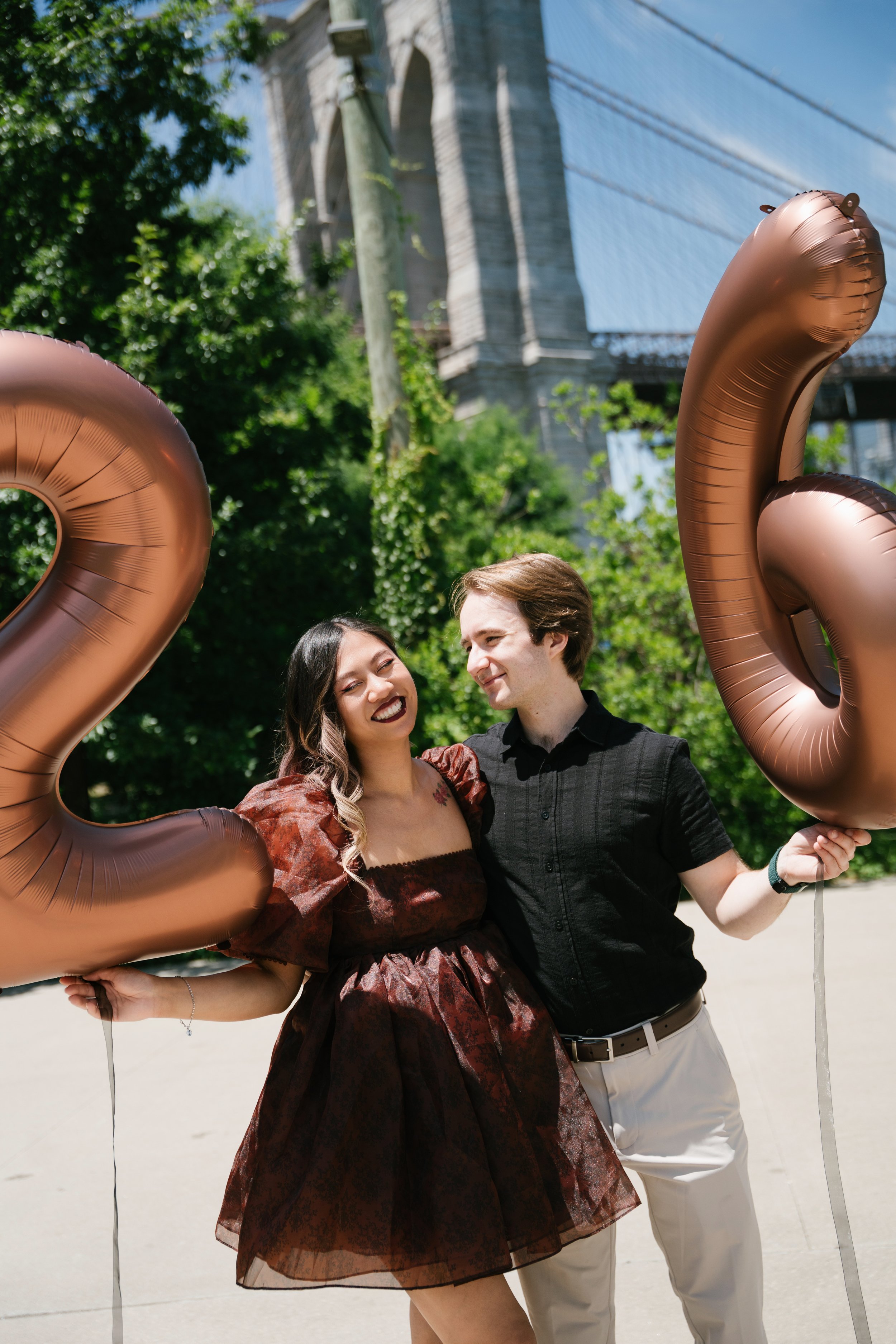 A happy young couple smiling and holding large rose gold balloons in the shape of the number 20, standing outdoors with a bridge in the background and trees around.