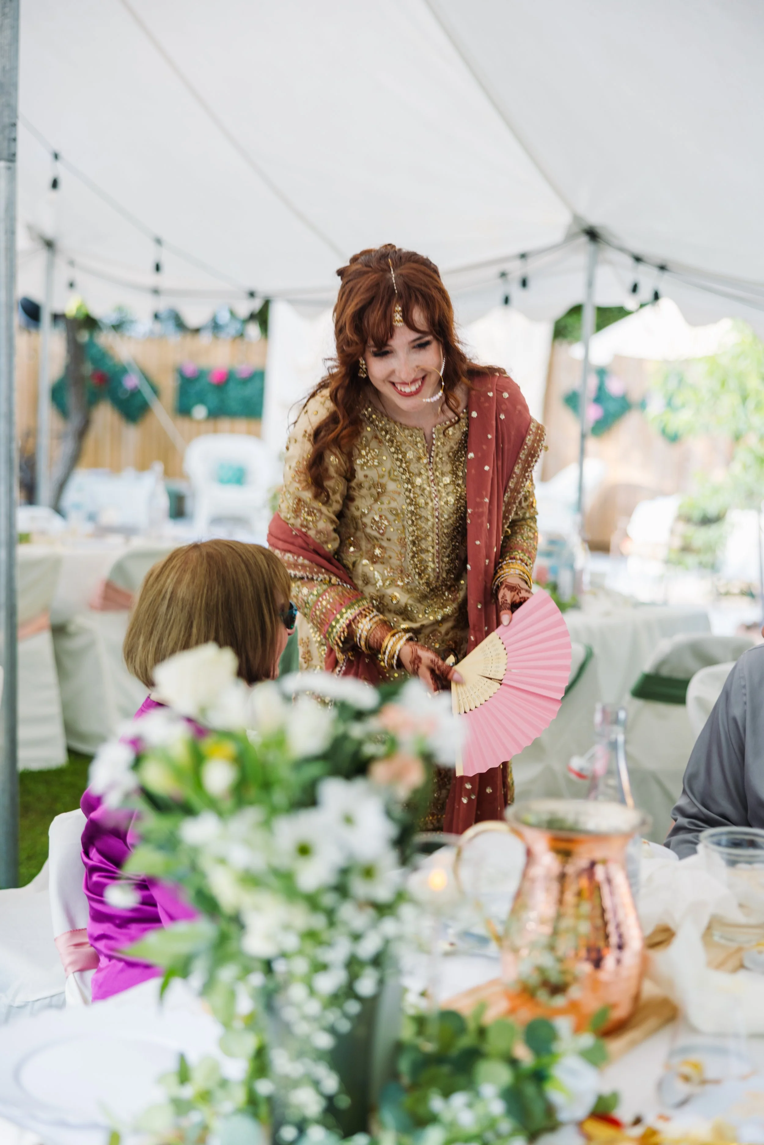 A woman in traditional South Asian attire smiling and serving food at a celebration under a white tent.