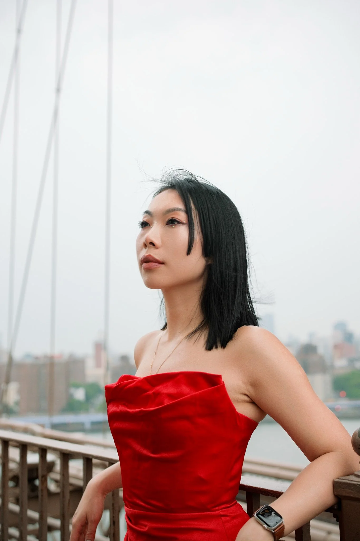 A woman with black hair wearing a red strapless dress and a smartwatch on her left wrist, standing on a balcony with city buildings and a cloudy sky in the background.