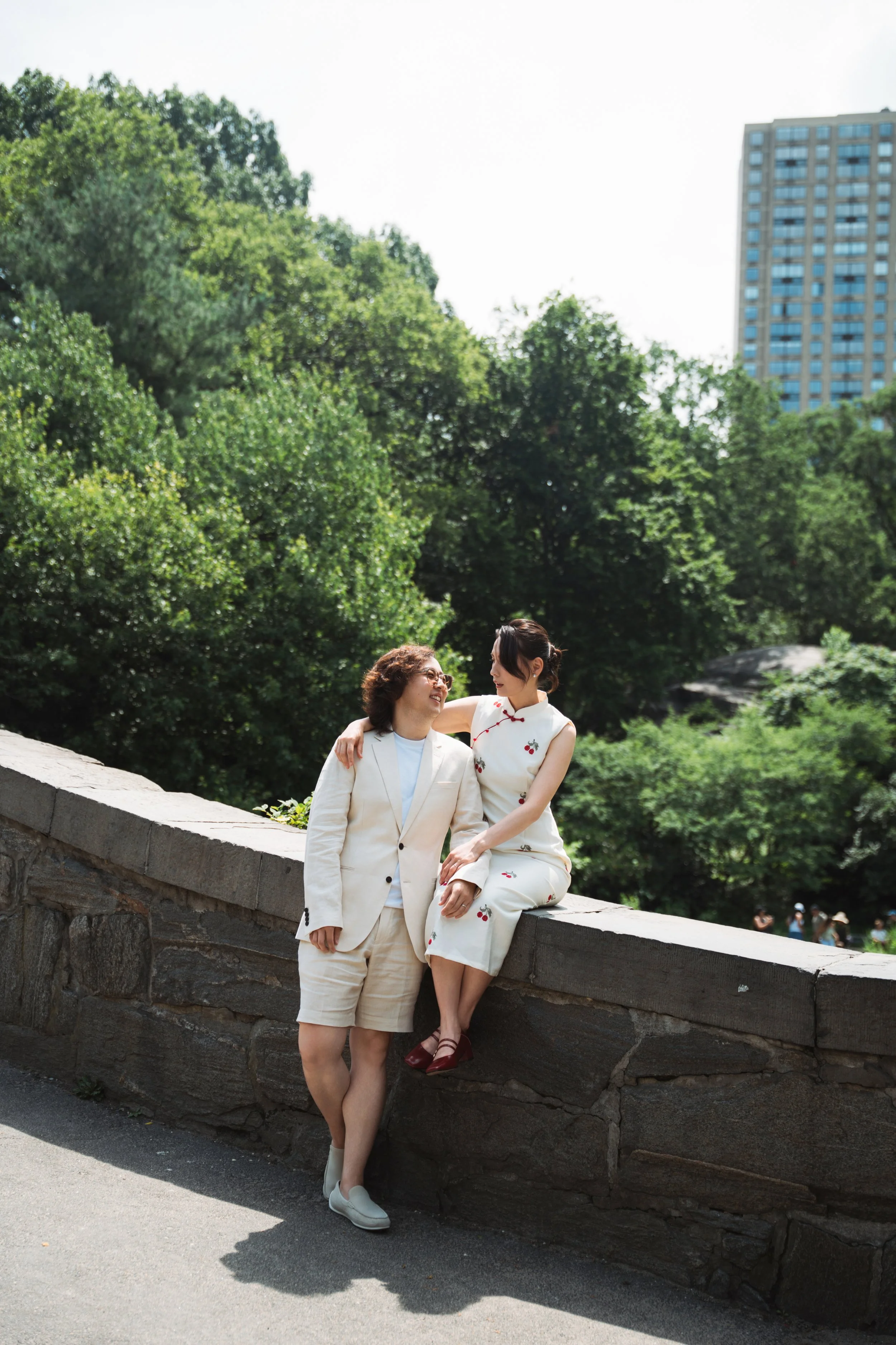 Two women, one standing and one sitting on a stone ledge, smiling and looking at each other in a park with green trees and a tall building in the background.