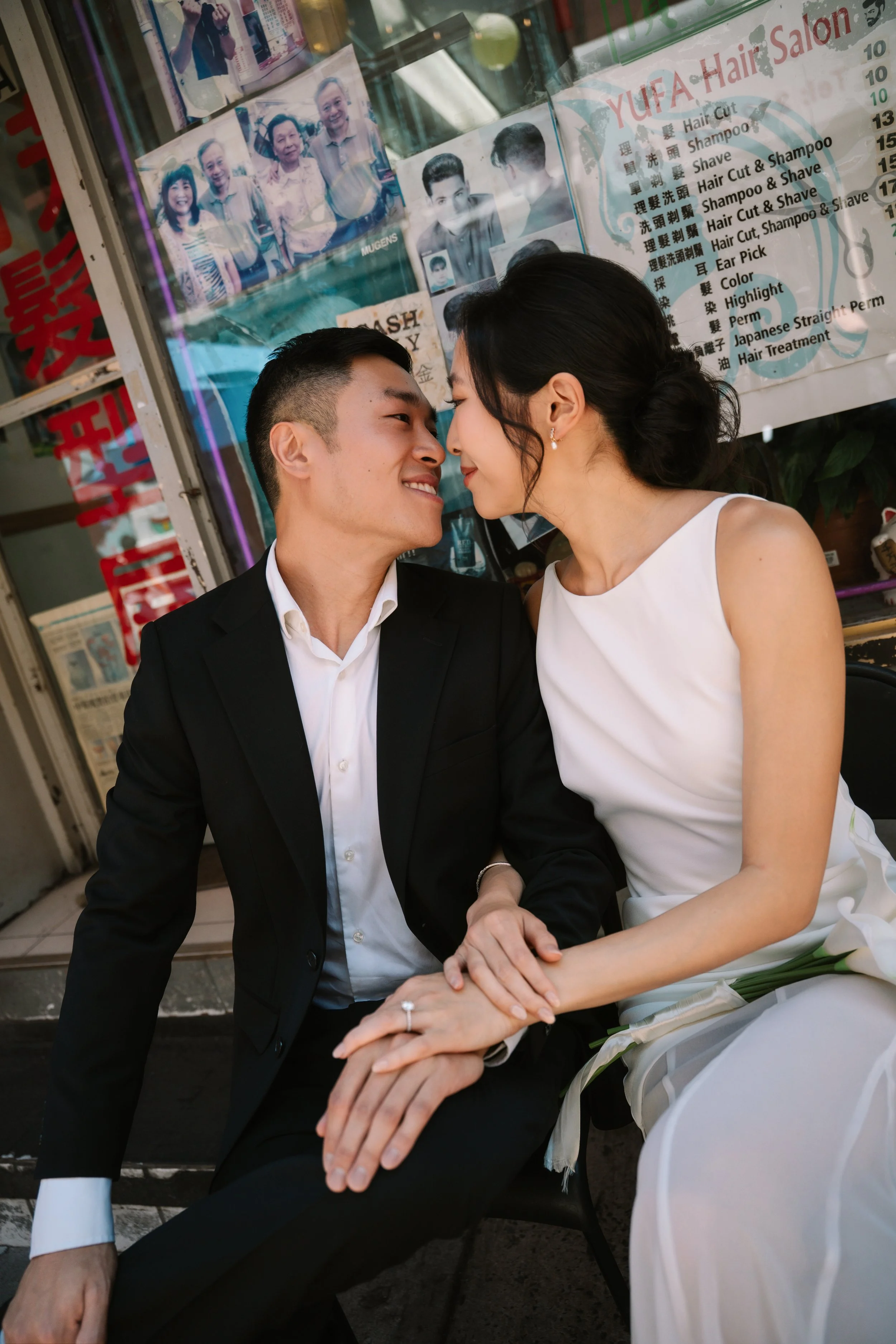 A happy couple, dressed in wedding attire, sit close together on a city sidewalk, leaning in with foreheads touching, sharing an intimate moment. Behind them is a glass window with photos and a sign listing various hair salon services.