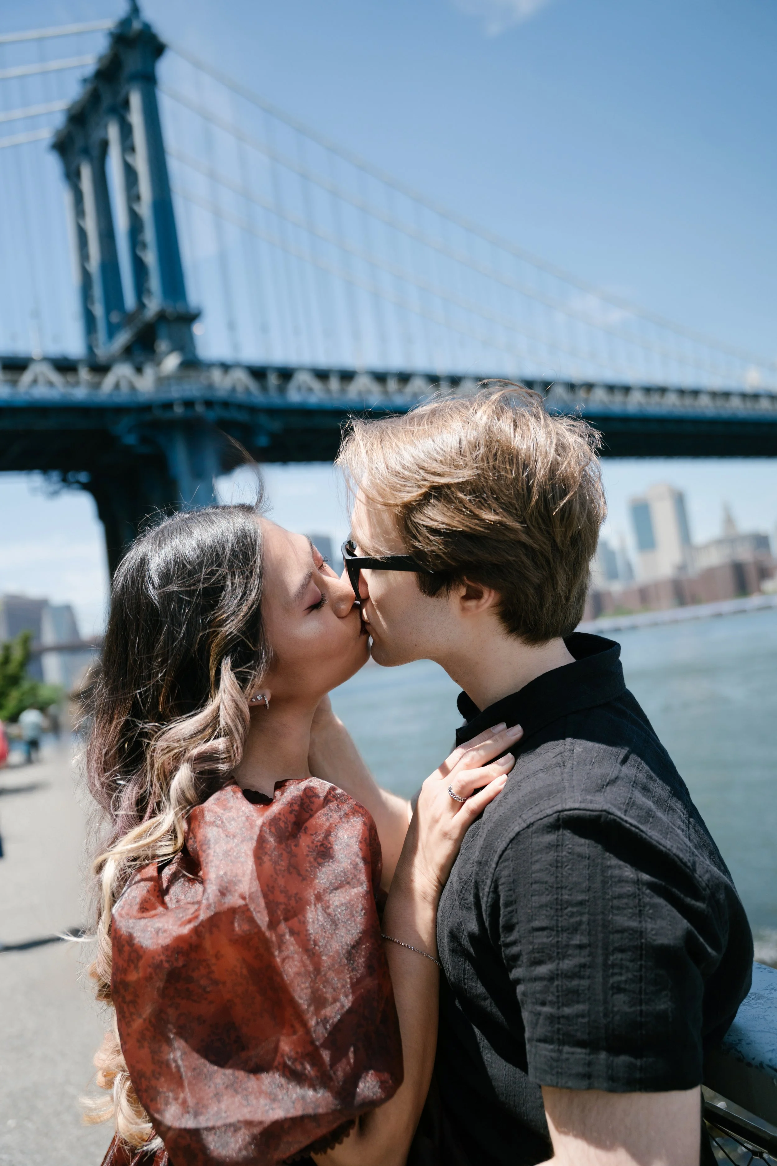 A couple kissing by a waterfront with a bridge in the background.