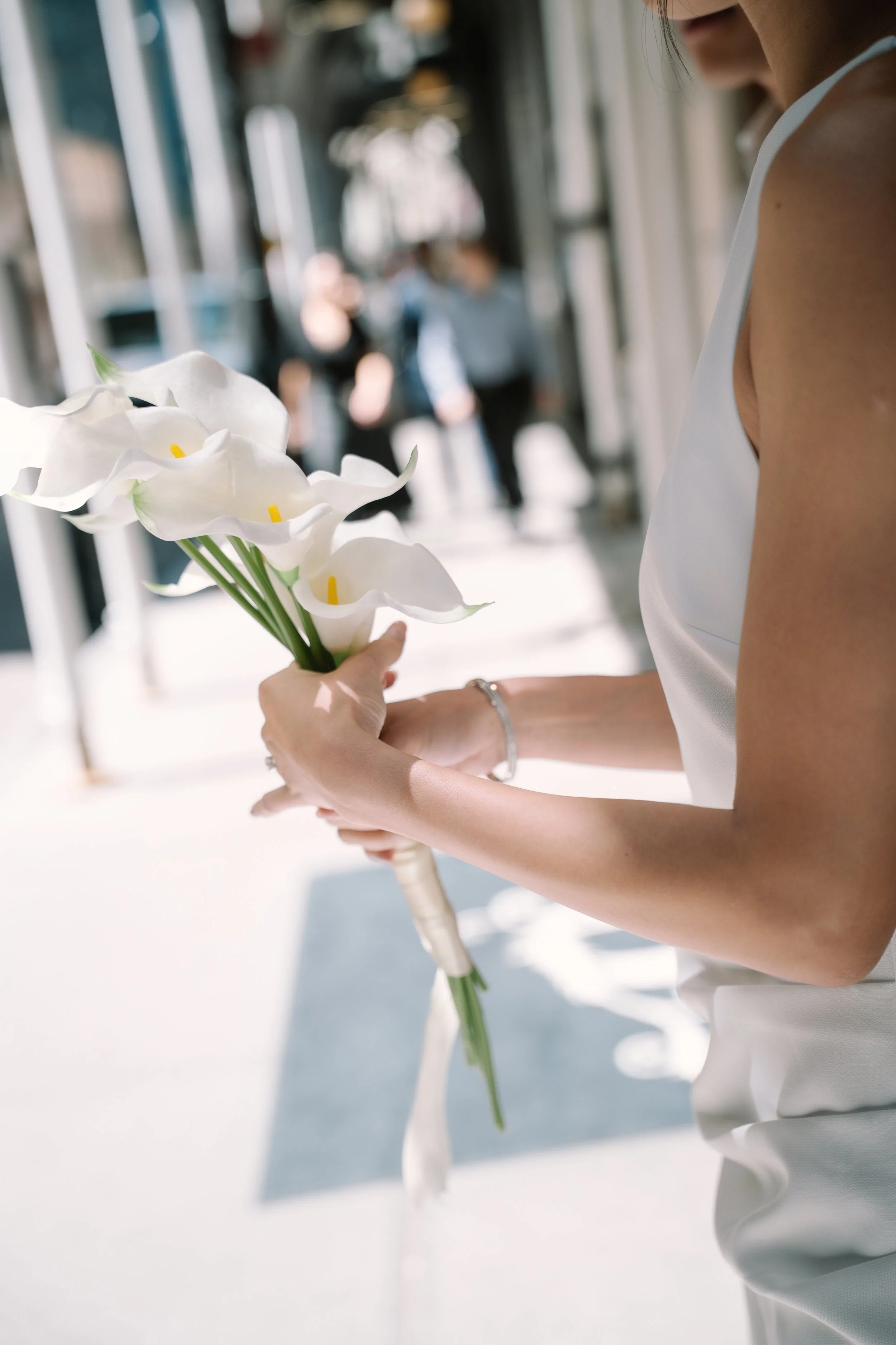 A woman dressed in white holding a bouquet of white calla lilies in an indoor setting with blurred people in the background.