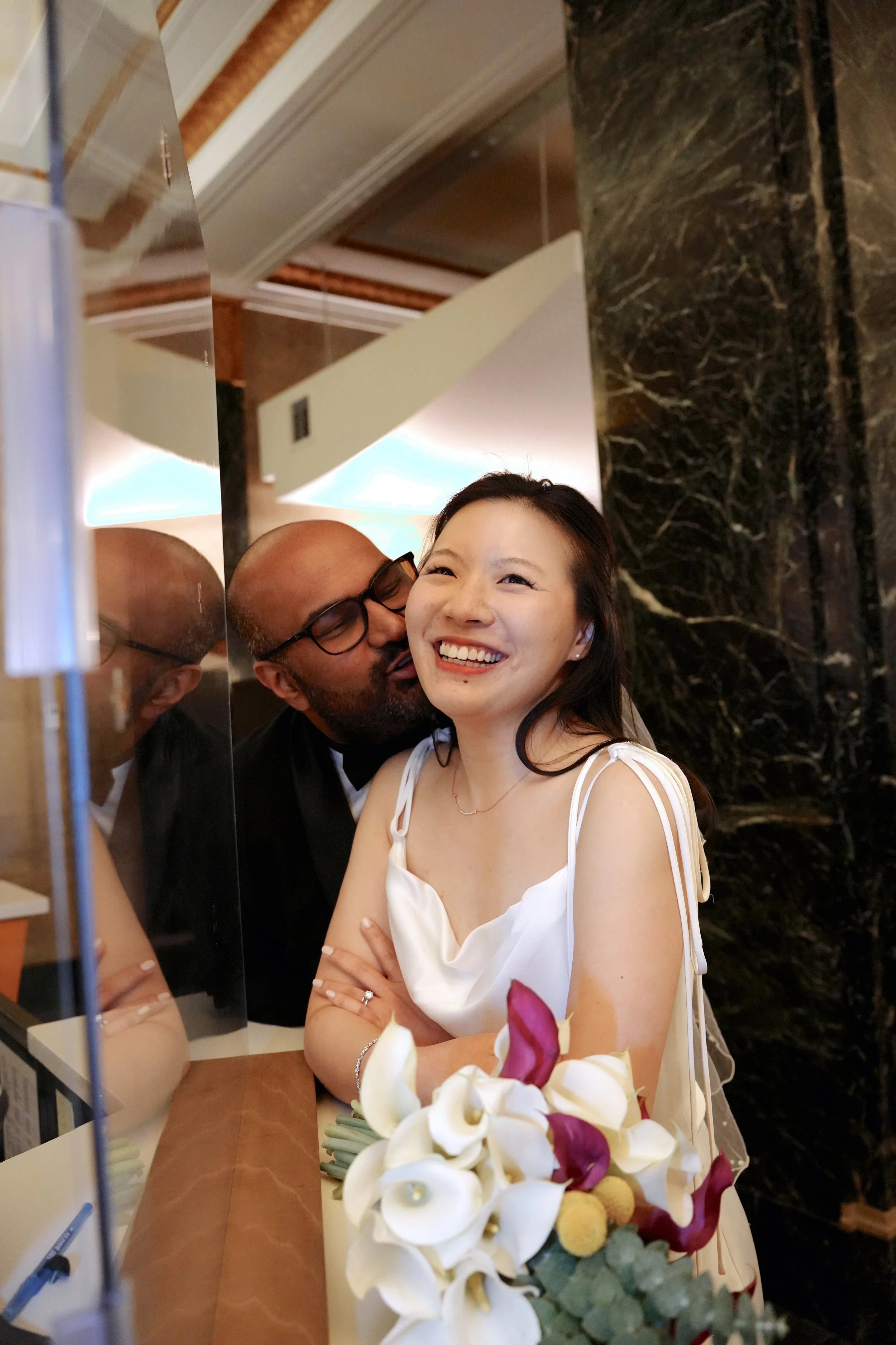 A man in glasses and a woman in a white dress smiling behind a decorative flower arrangement, seen through a mirror in a hotel lobby.