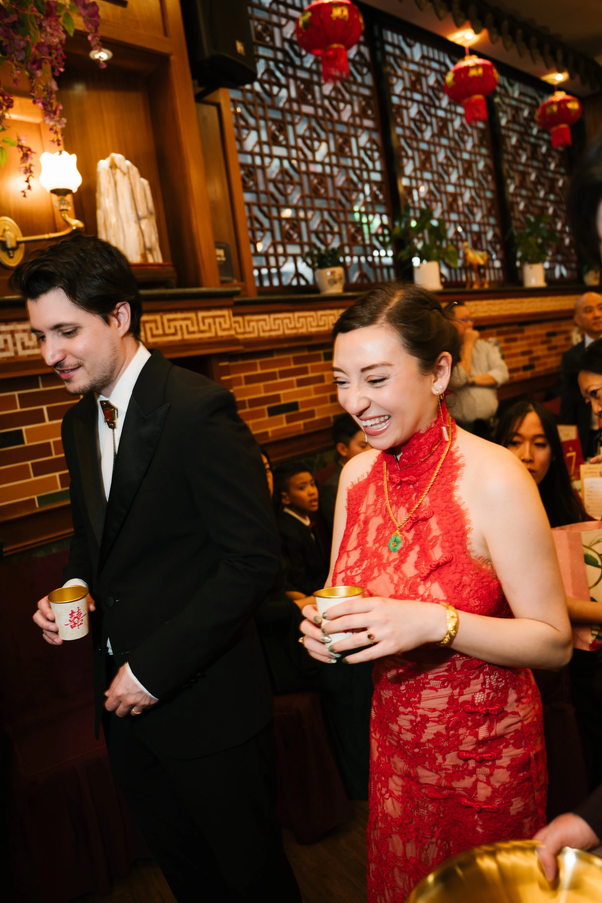 Two people dressed in formal attire, holding cups, participating in a celebration in a restaurant decorated with Chinese lanterns and traditional decor.