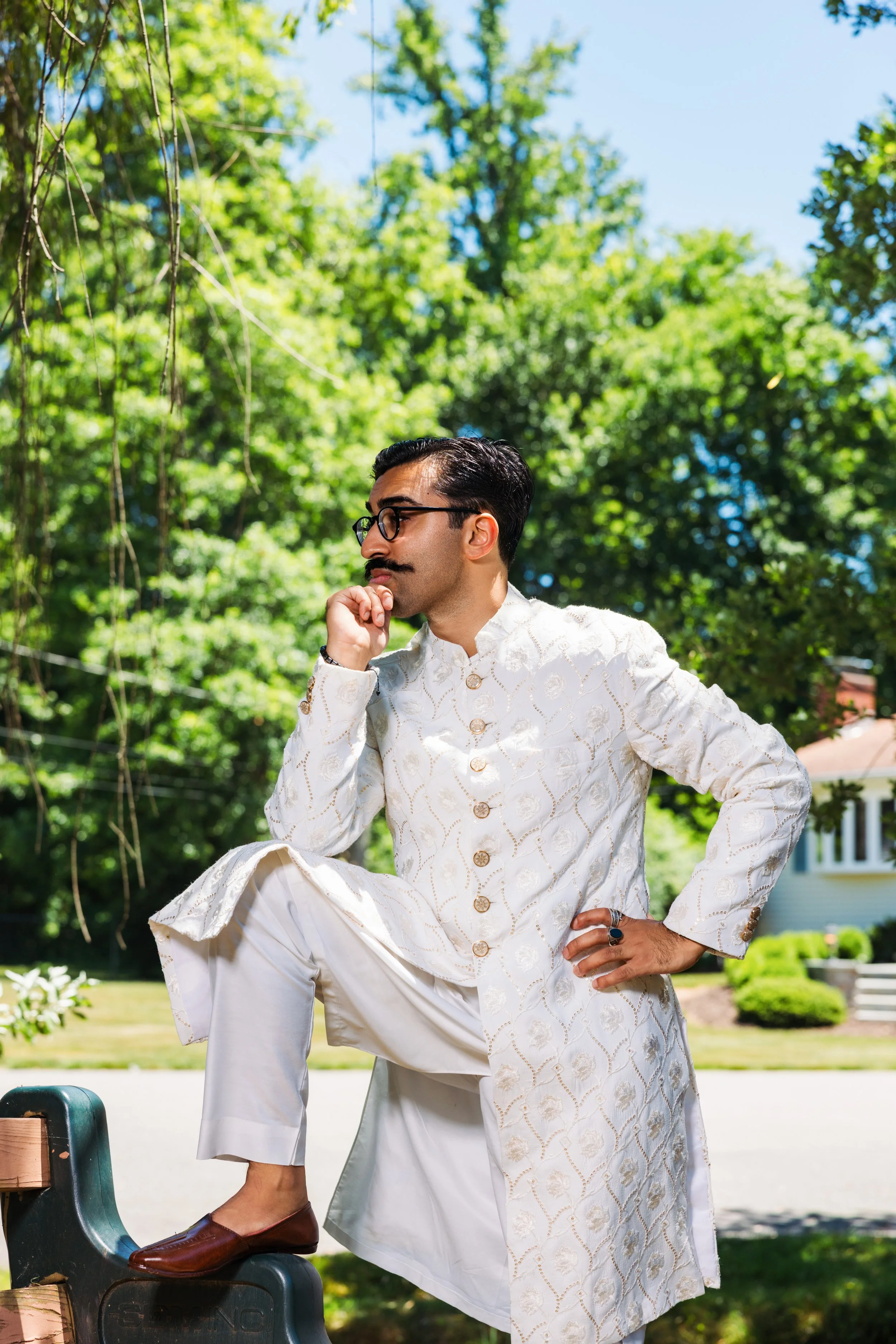 A man dressed in traditional cream-colored Indian attire, wearing glasses, gold jewelry, and a ring, is sitting on a park bench with one foot on a step, surrounded by green trees and a house in the background.
