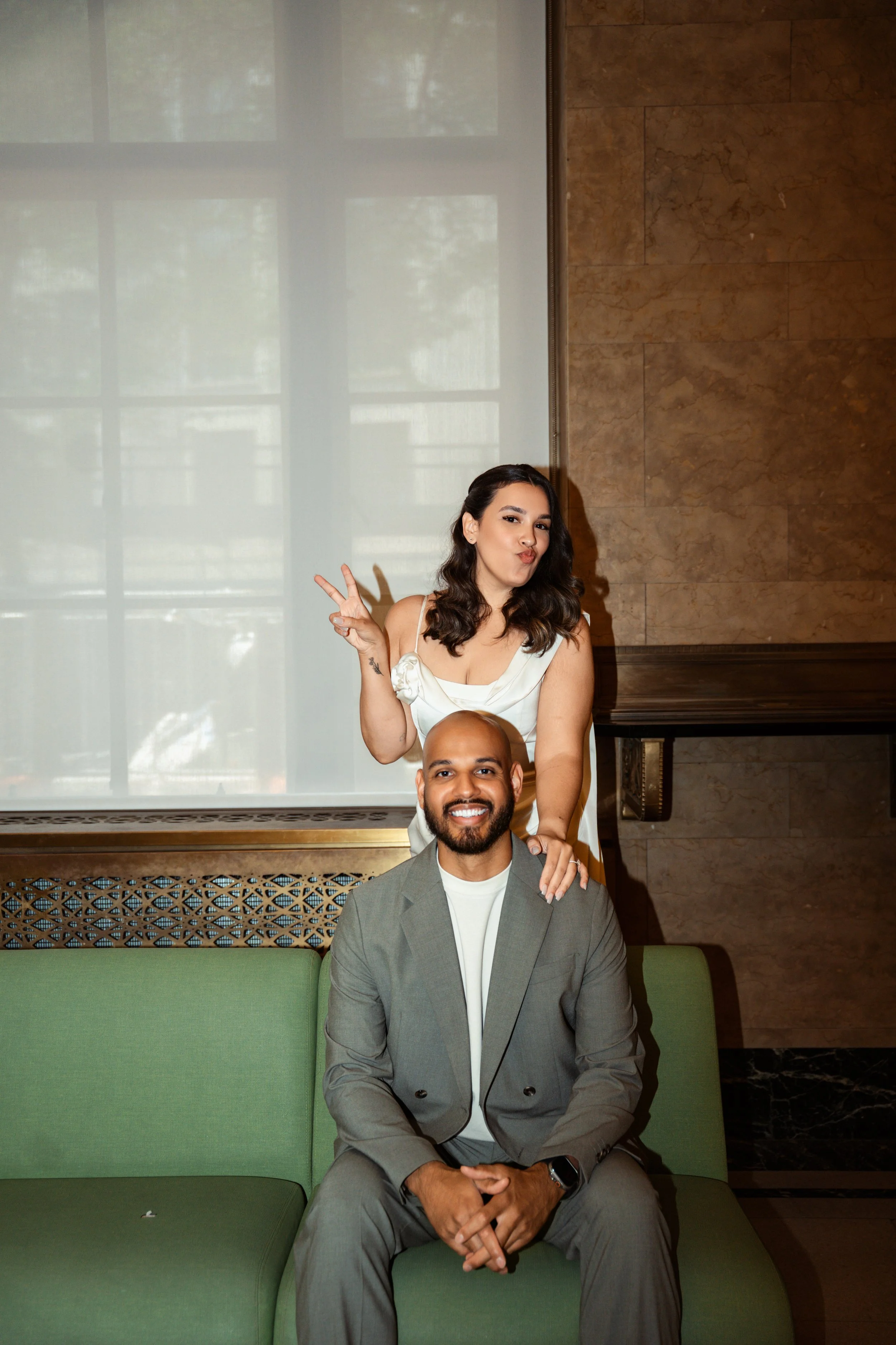 A woman is making a peace sign and a playful face, sitting behind a man in a grey suit who is smiling and seated in front of a green couch in an indoor setting with a marble wall and large windows.