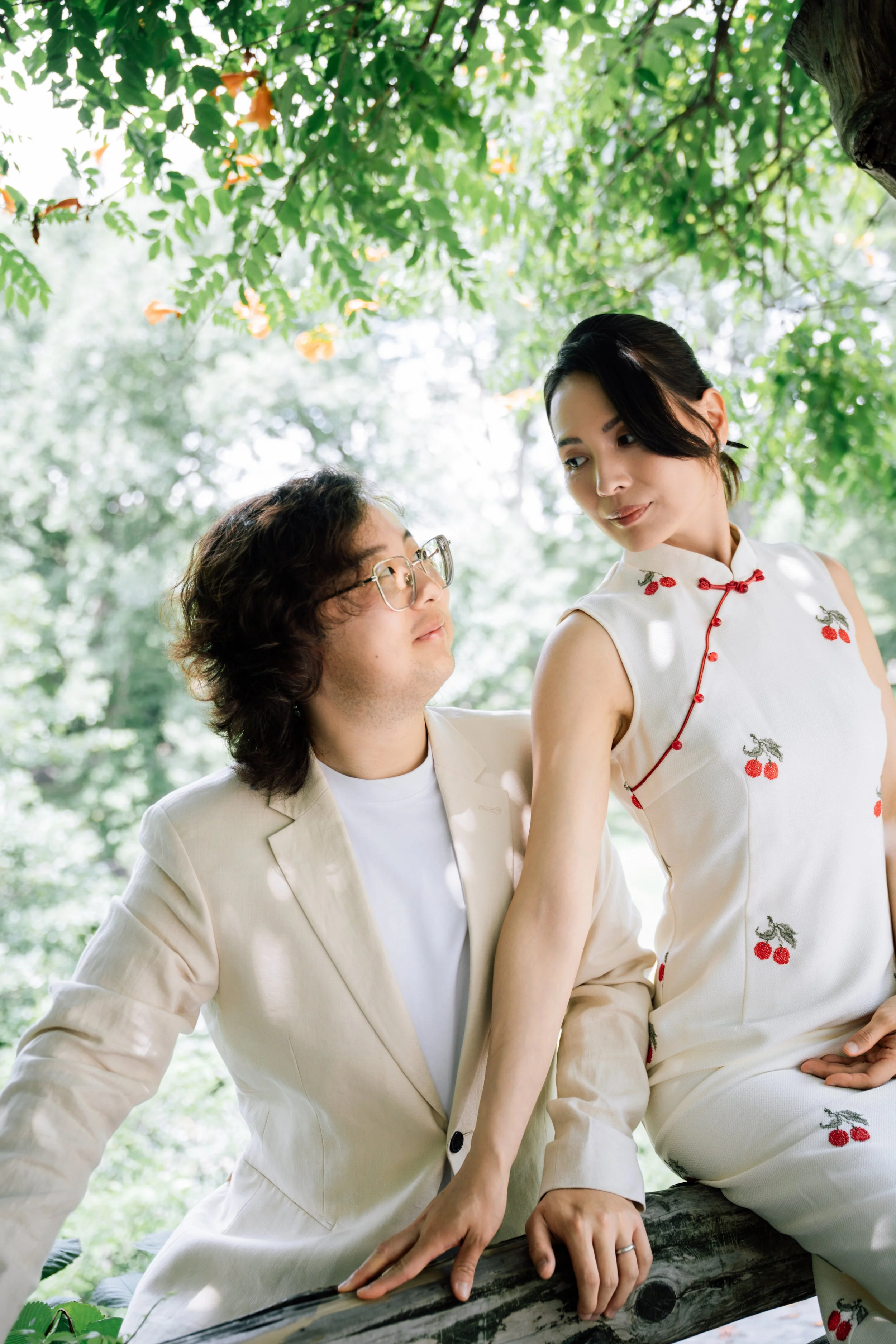 A man and woman sitting outdoors under a leafy tree, engaged in conversation. The woman is wearing a white dress with cherry embroidery, and the man is in a light-colored blazer. They are seated on a log surrounded by green foliage.