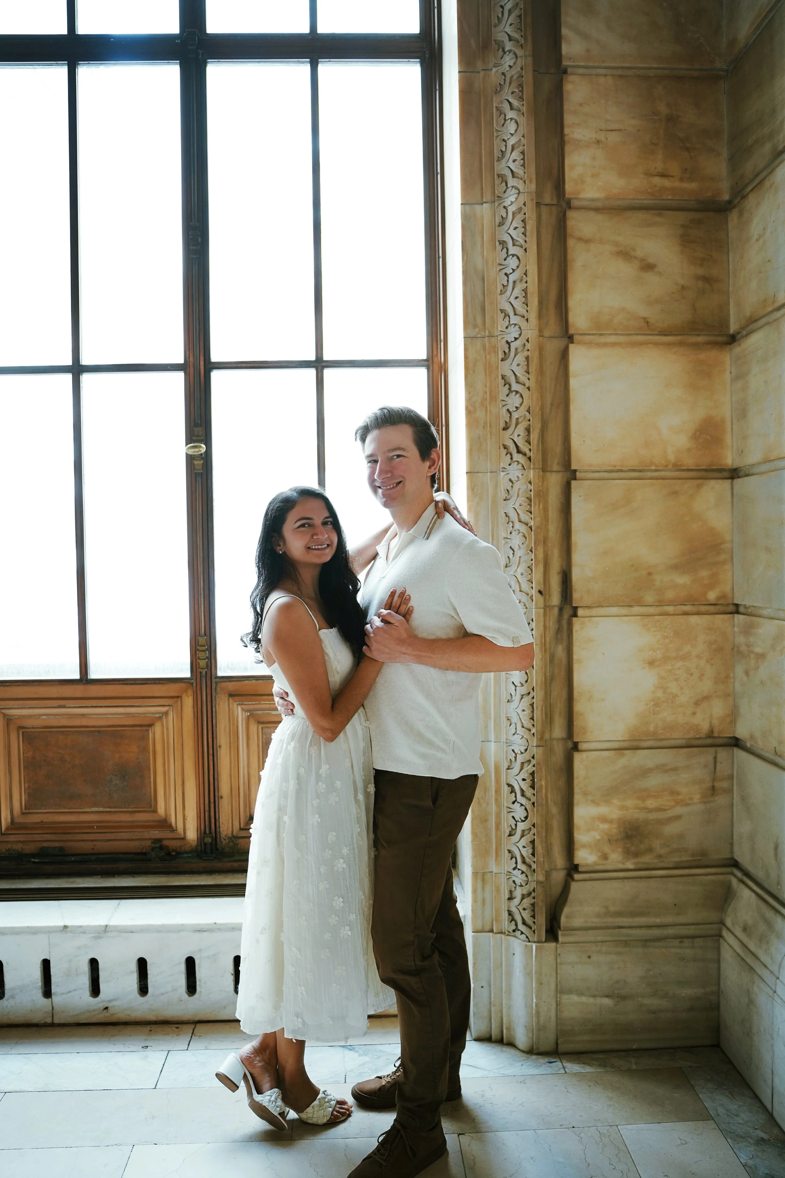 A smiling couple embraces and poses inside a historic building with large windows and ornate stonework.