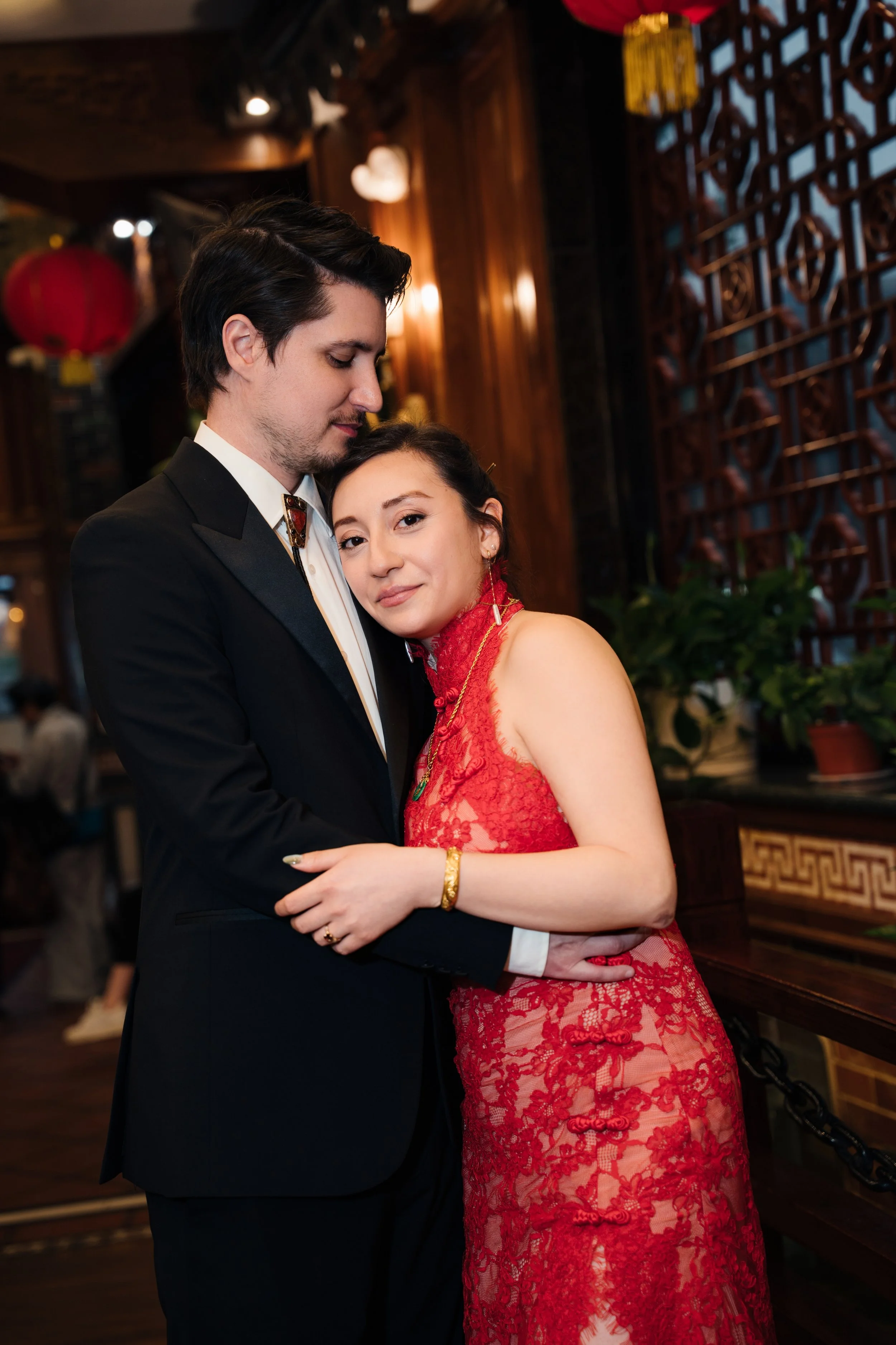 A young man in a black tuxedo and a young woman in a red lace dress are standing close together, embracing, in a warmly lit restaurant decorated with wooden panels, Chinese lanterns, and traditional decor.