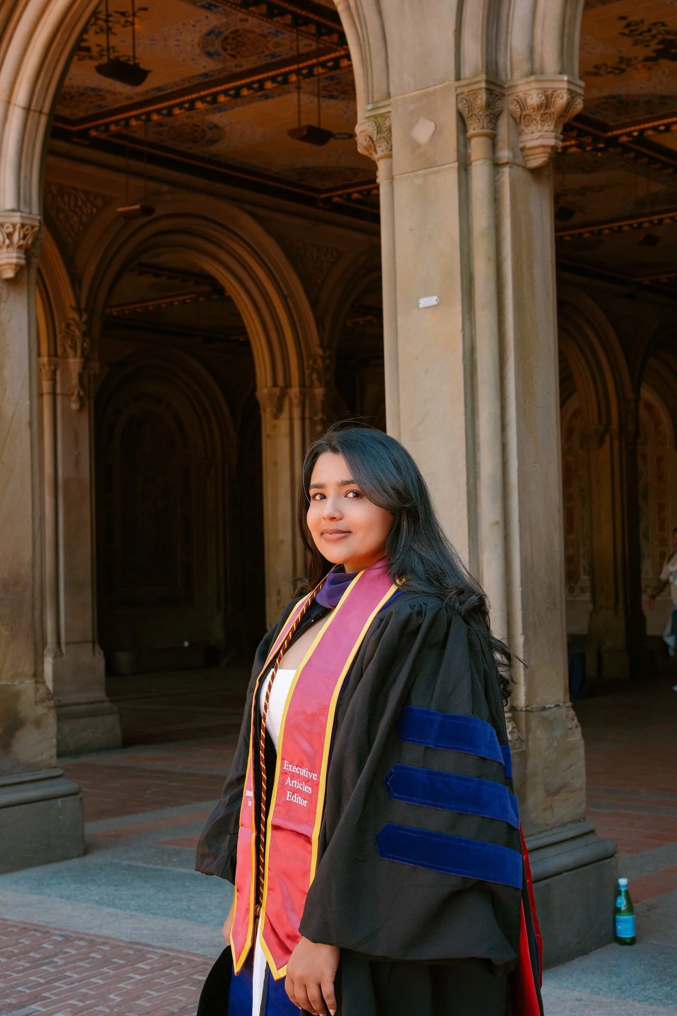 A young woman in a graduation cap and gown standing outdoors in front of an ornate stone building.
