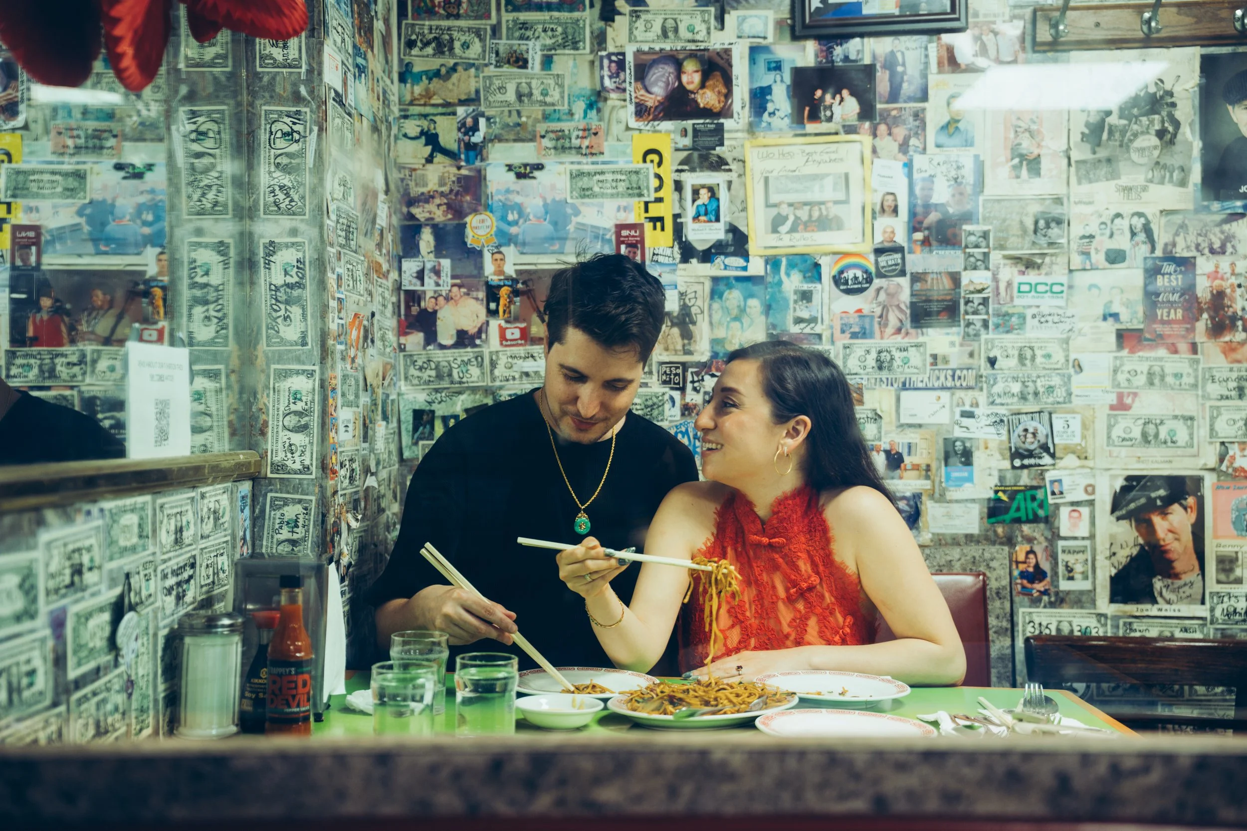 A young man and woman dining together in a restaurant decorated with magazine clippings and photographs on the wall. The woman is wearing a red lace dress, and they are sharing a meal with chopsticks, with plates of noodles and glasses of drinks in f