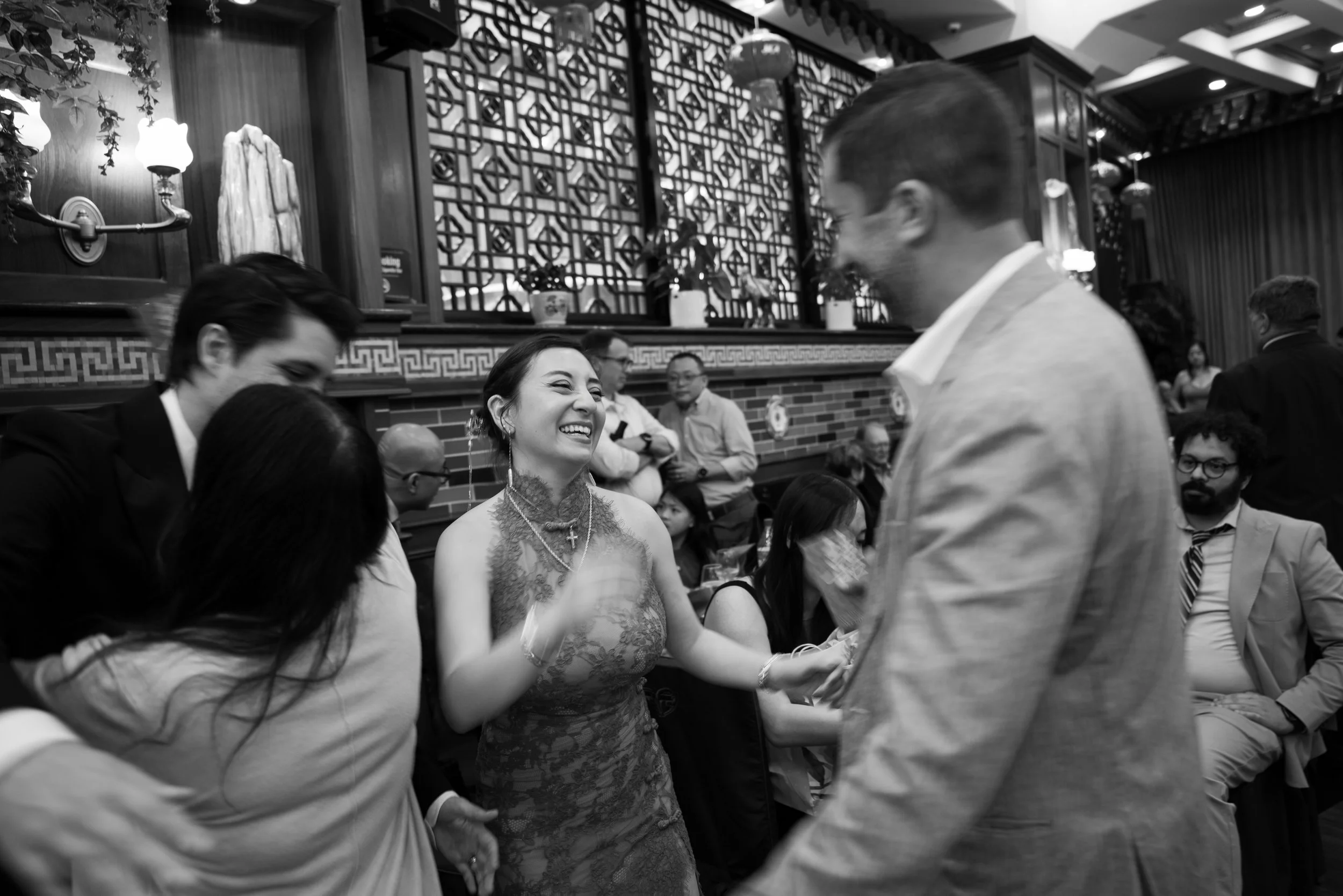 A woman with dark hair and lace dress smiling and holding hands with someone at a social gathering in a restaurant with Asian decorations.