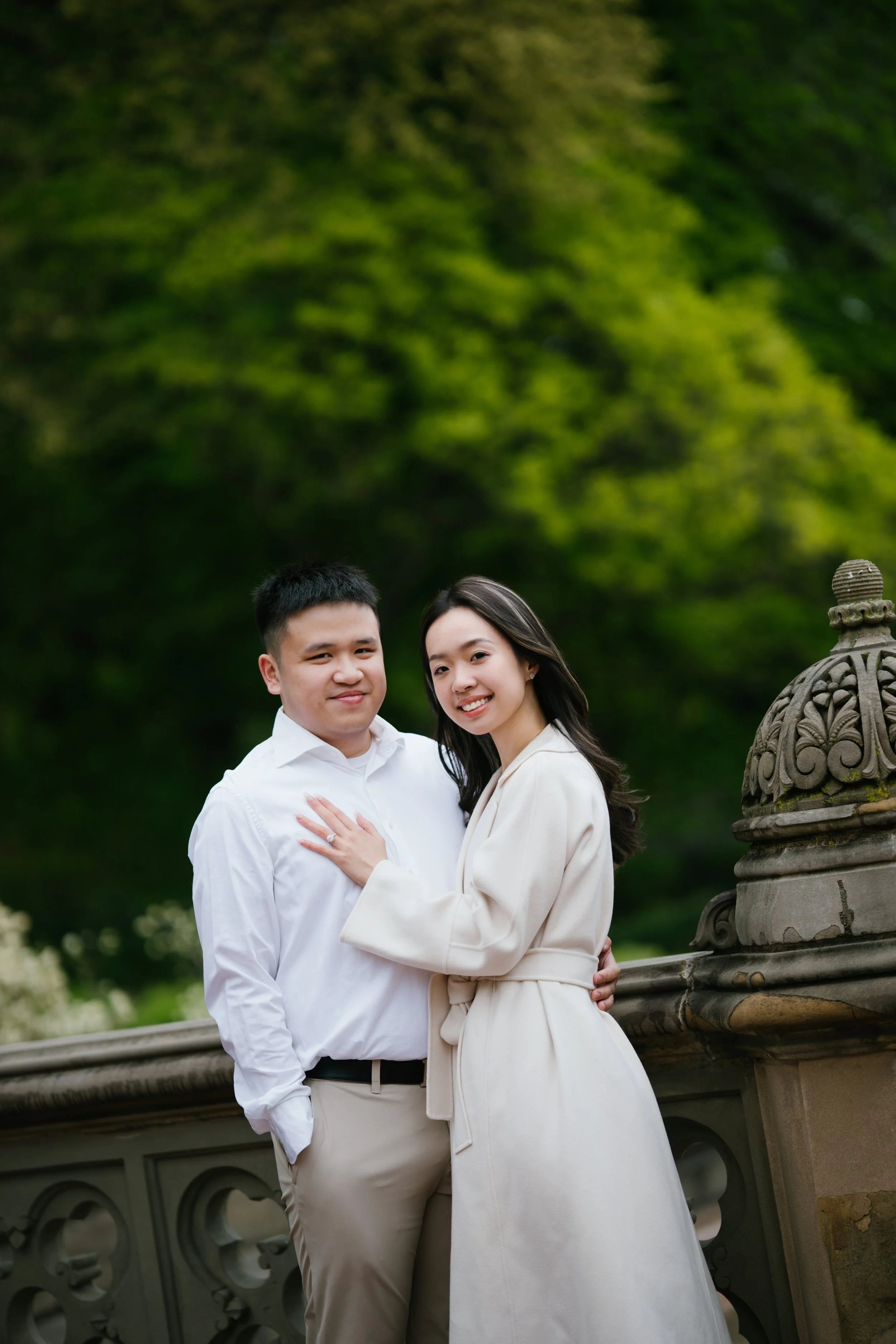 A couple embracing outdoors, standing on a stone bridge with greenery in the background.