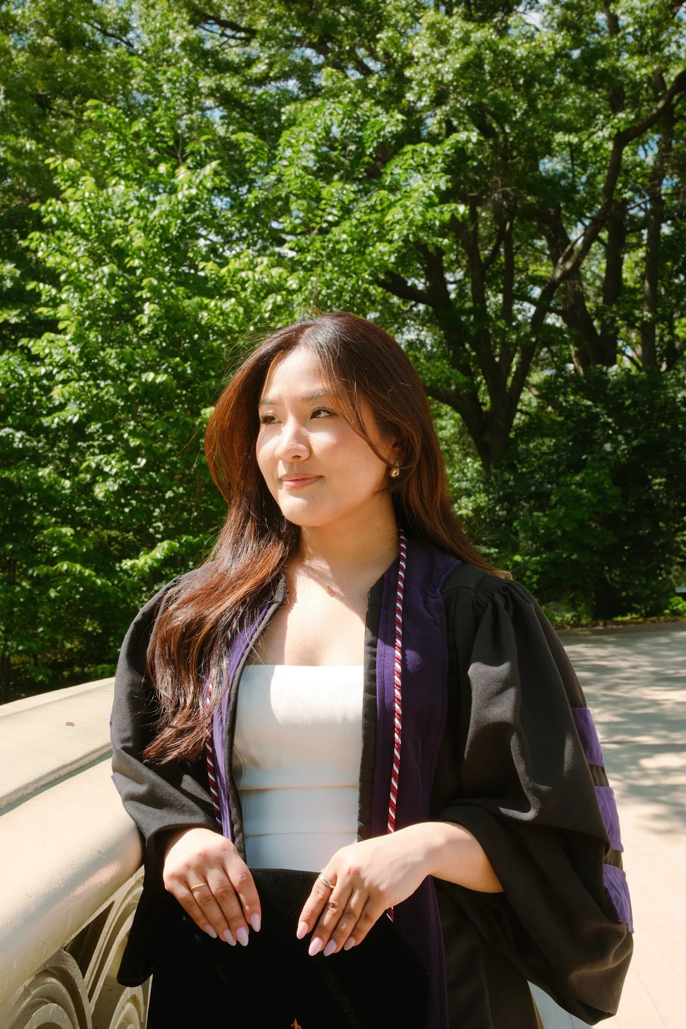 Young woman with long brown hair, wearing a white top and a black graduation gown with purple trim, standing outdoors near a beige railing, with green trees in the background on a sunny day.