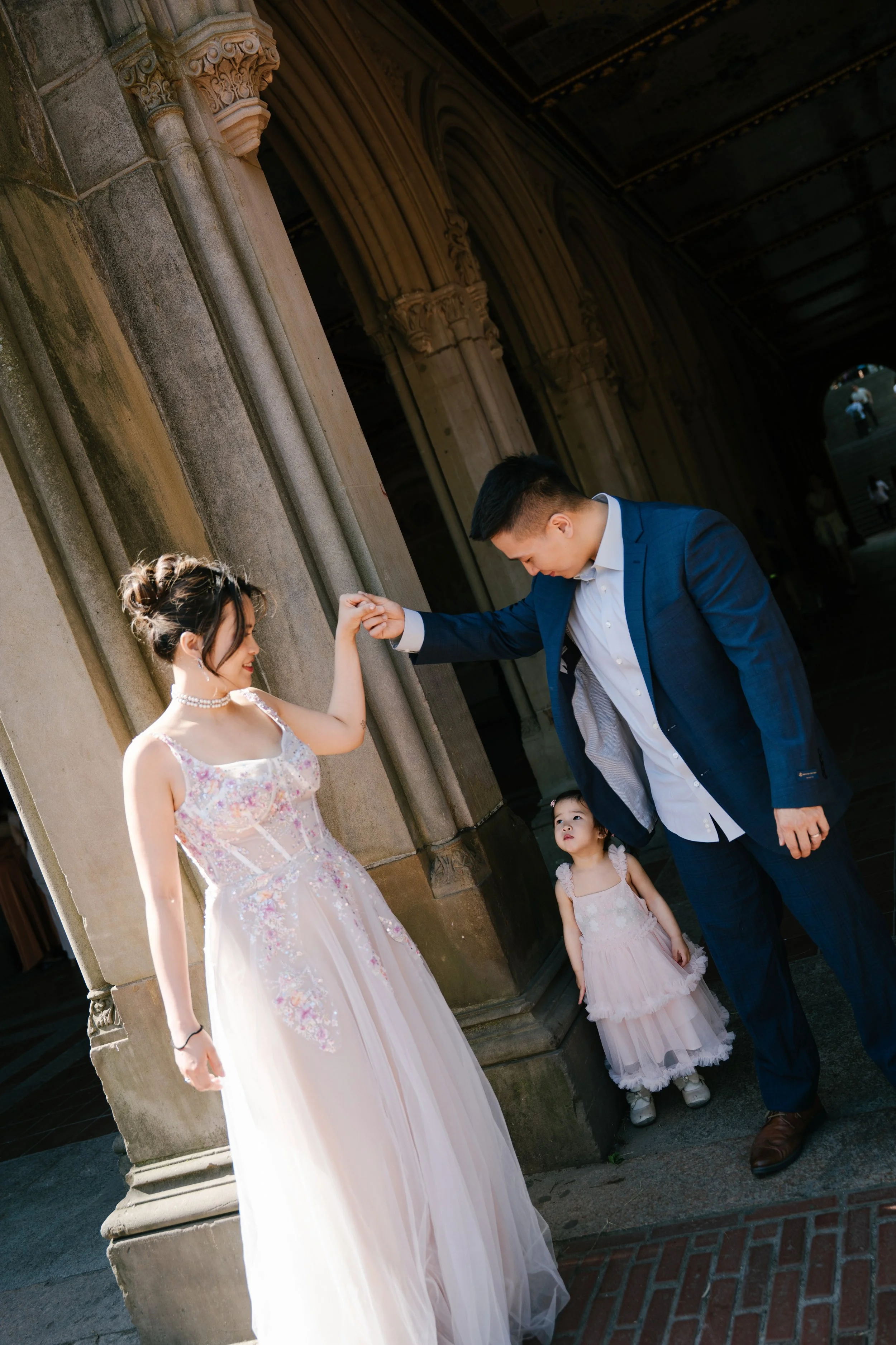A young woman in a floral dress and pearl necklace is arm wrestling with a young man in a blue suit and white shirt. A little girl in a pink dress looks on, standing near them outside a stone building with ornate columns.