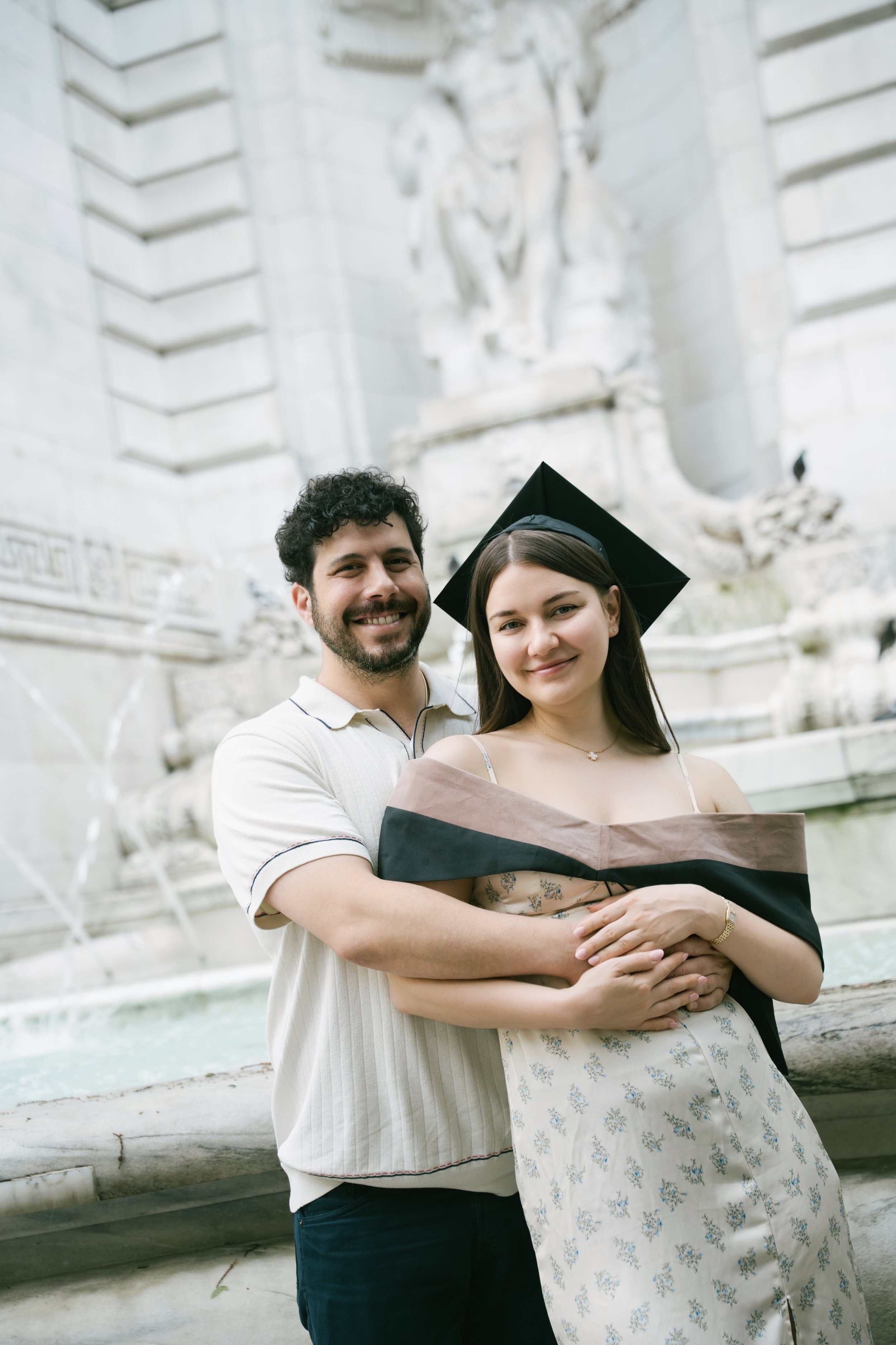 A young woman and man smiling, with the woman in a graduation cap and gown and the man hugging her from behind, standing in front of a large, white stone monument or fountain.