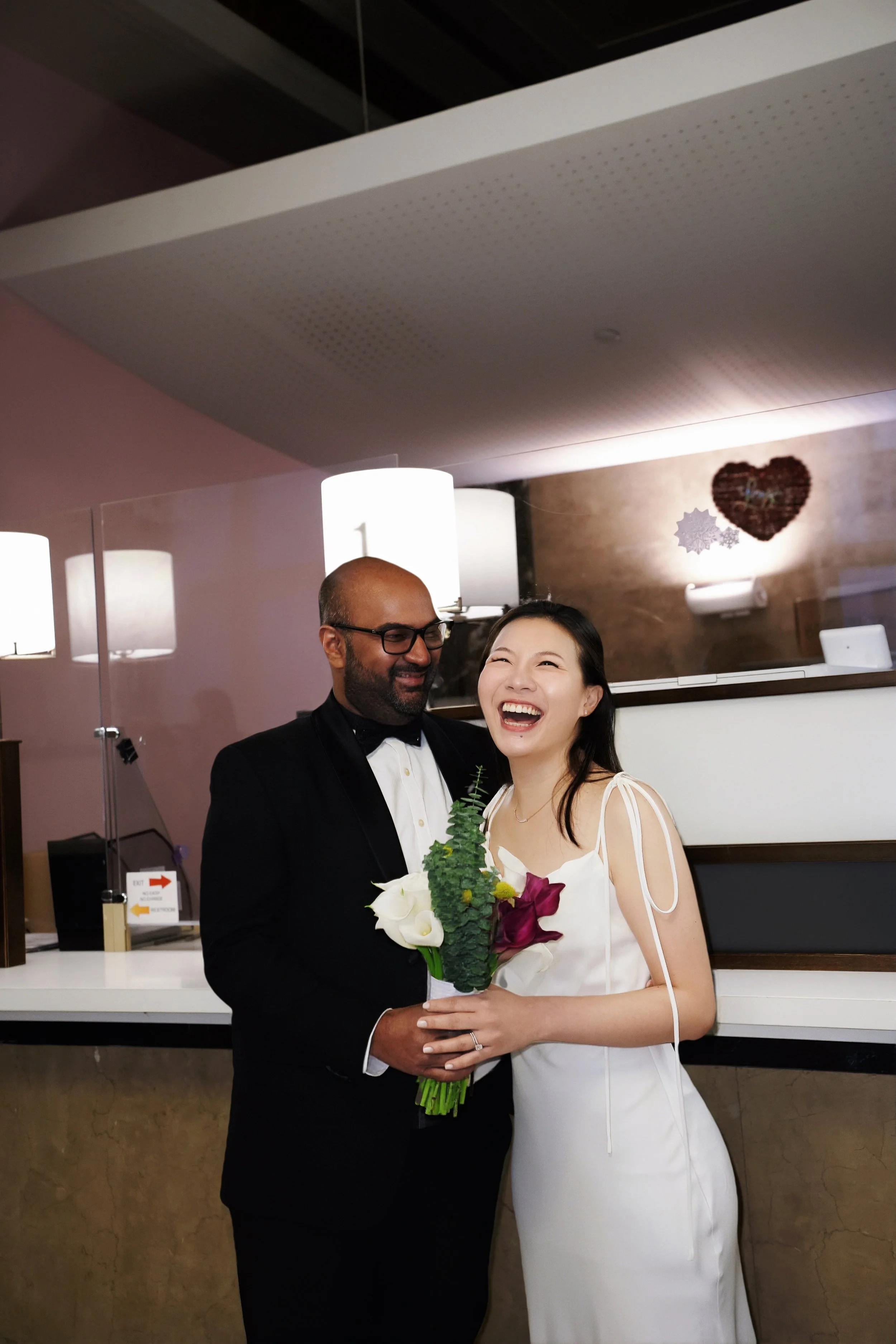 A man in a tuxedo with glasses and a woman in a white dress are smiling and holding a bouquet of white and purple flowers in a hotel or event hall with warm lighting and wall decor that includes a heart-shaped decoration.