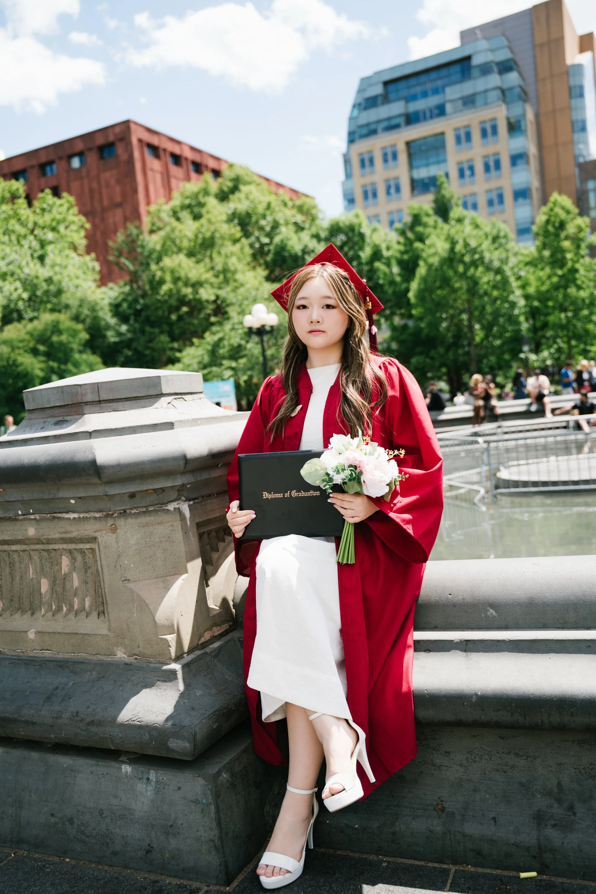 Young woman in a red graduation gown and cap holding a diploma and flowers, standing on a city street near a stone structure with buildings and trees in the background.