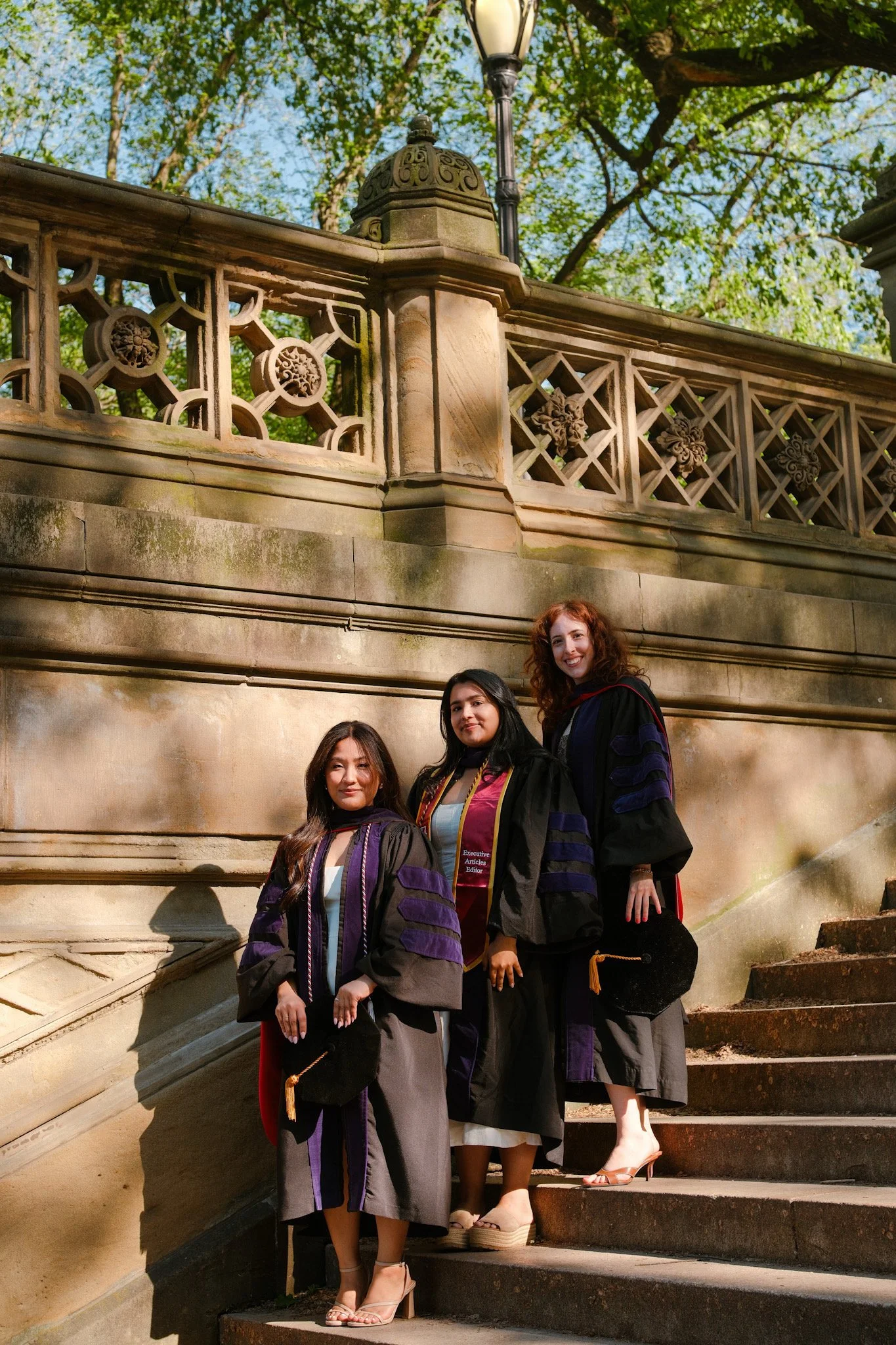 Three women in graduation caps and gowns standing on the steps of a historic building with ornate stone railings and lush green trees in the background.