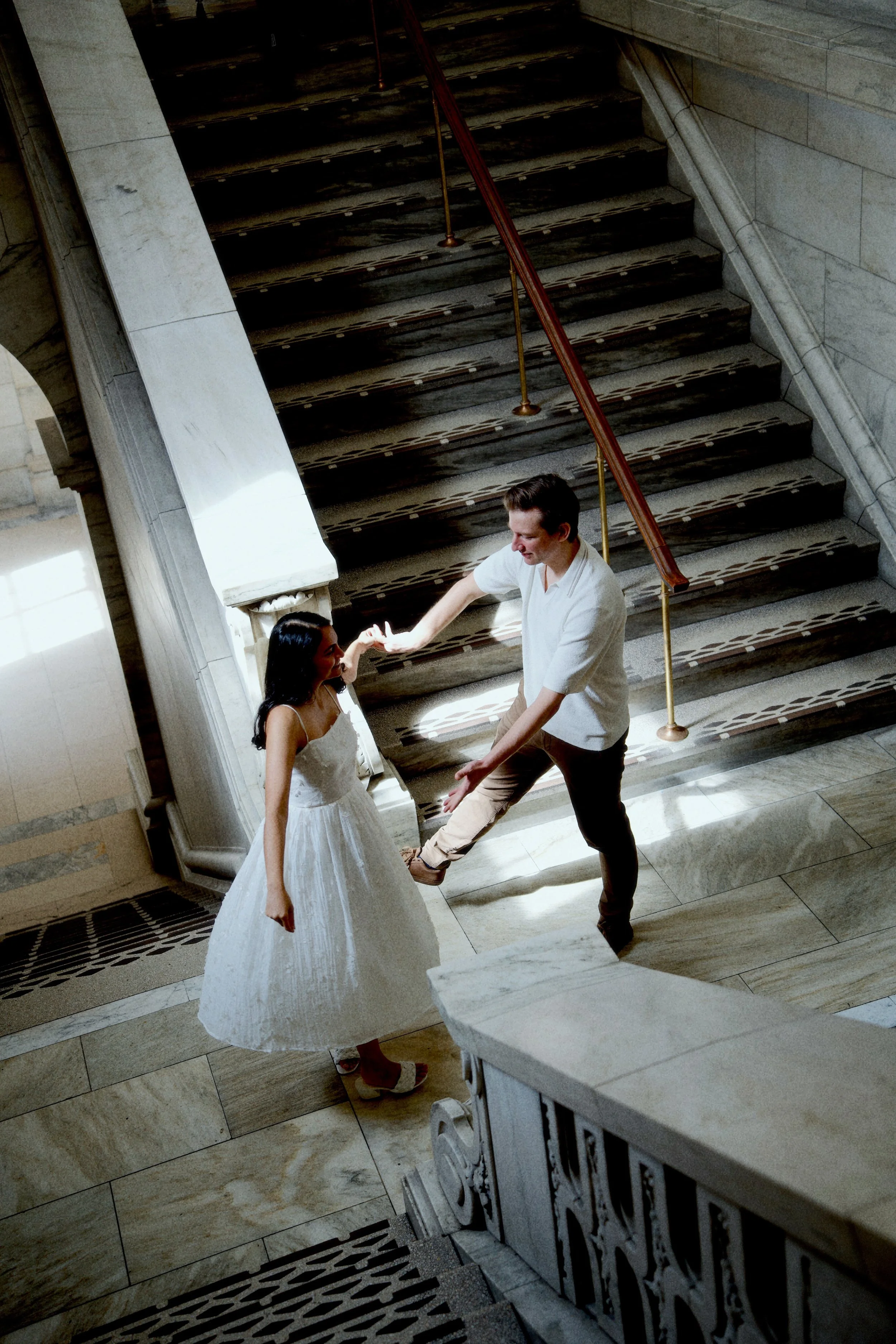 A man and woman on a grand staircase, with the man helping the woman with her shoe. The woman is in a white dress, and the man is in a white shirt and brown pants. The staircase has dark steps and a red handrail, with marble walls surrounding it.