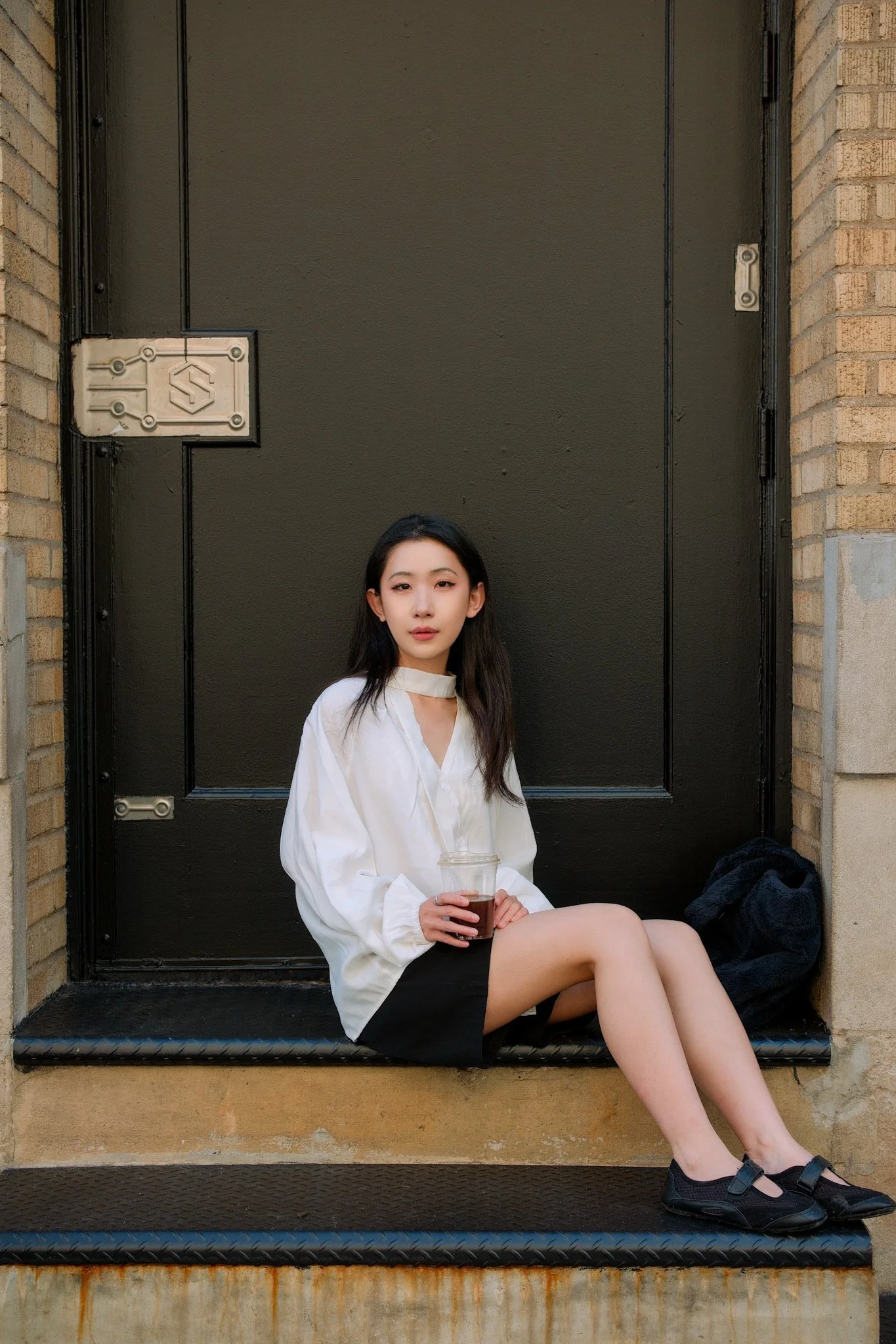 A young woman sitting on a black step in front of a black door, holding a glass of a dark beverage, wearing a white long sleeve shirt, black shorts, and black sandals.