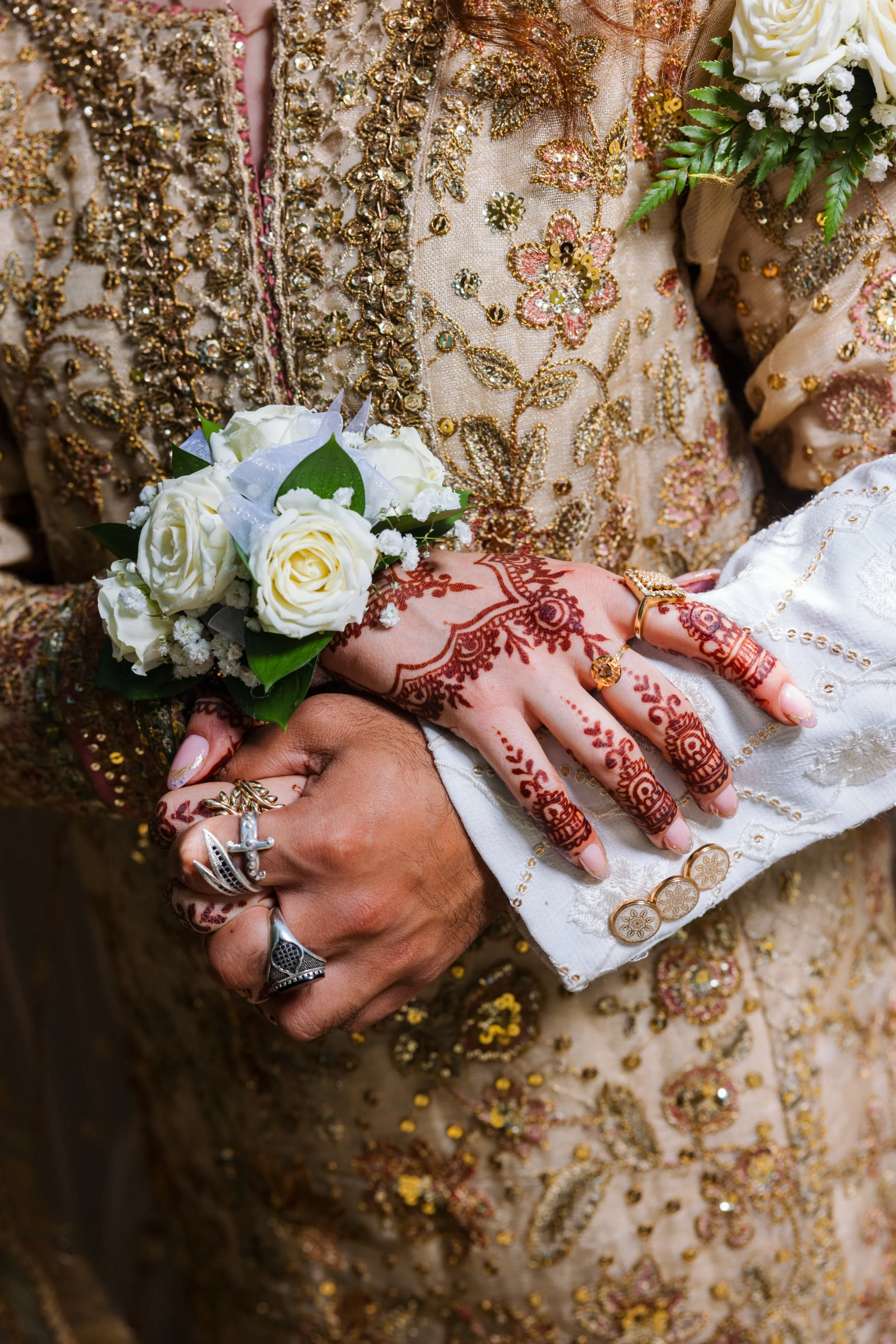 Close-up of a couple's hands adorned with multiple rings, holding a white floral bouquet. The hands are covered in intricate henna designs. Both individuals are wearing traditional, ornate, gold and bead-embellished clothing, with one person wearing 