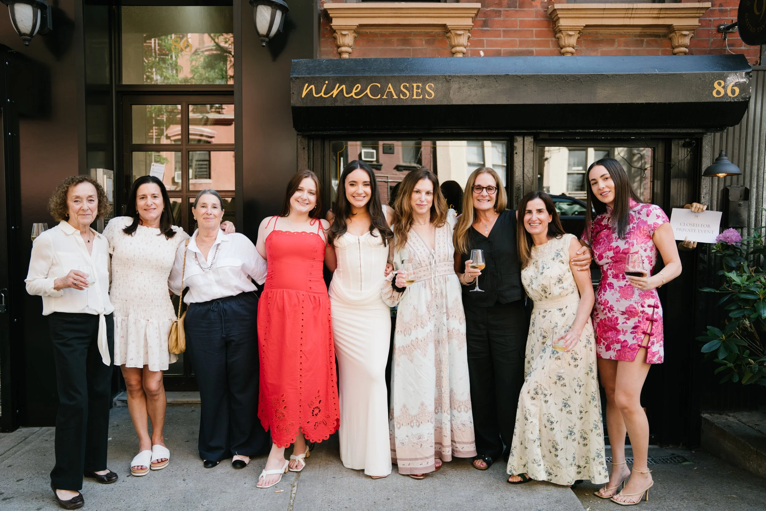 Group of nine women standing outside a restaurant called 'nineCASES,' smiling with some holding glasses of wine, dressed in summery dresses and casual attire.