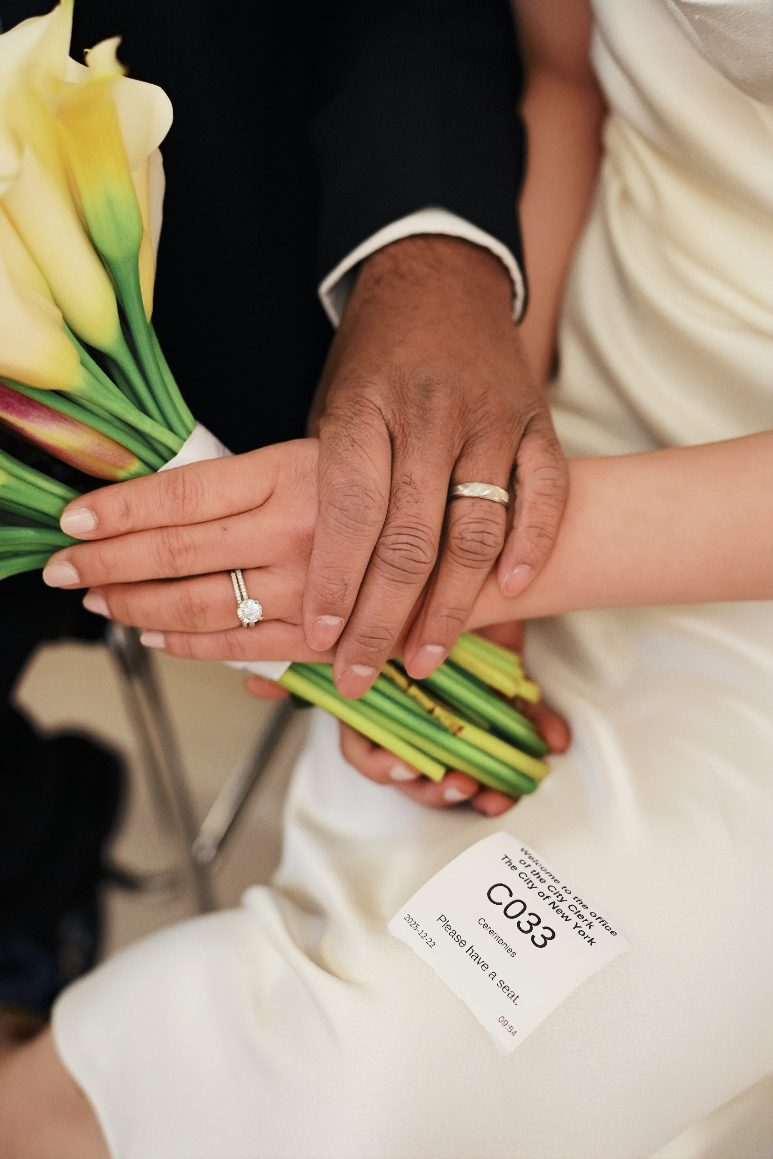 Close-up of two hands with wedding rings gently holding a bouquet of yellow calla lilies with green stems. One hand, with darker skin, is on top of the other, with lighter skin, holding a white event ticket with black text.