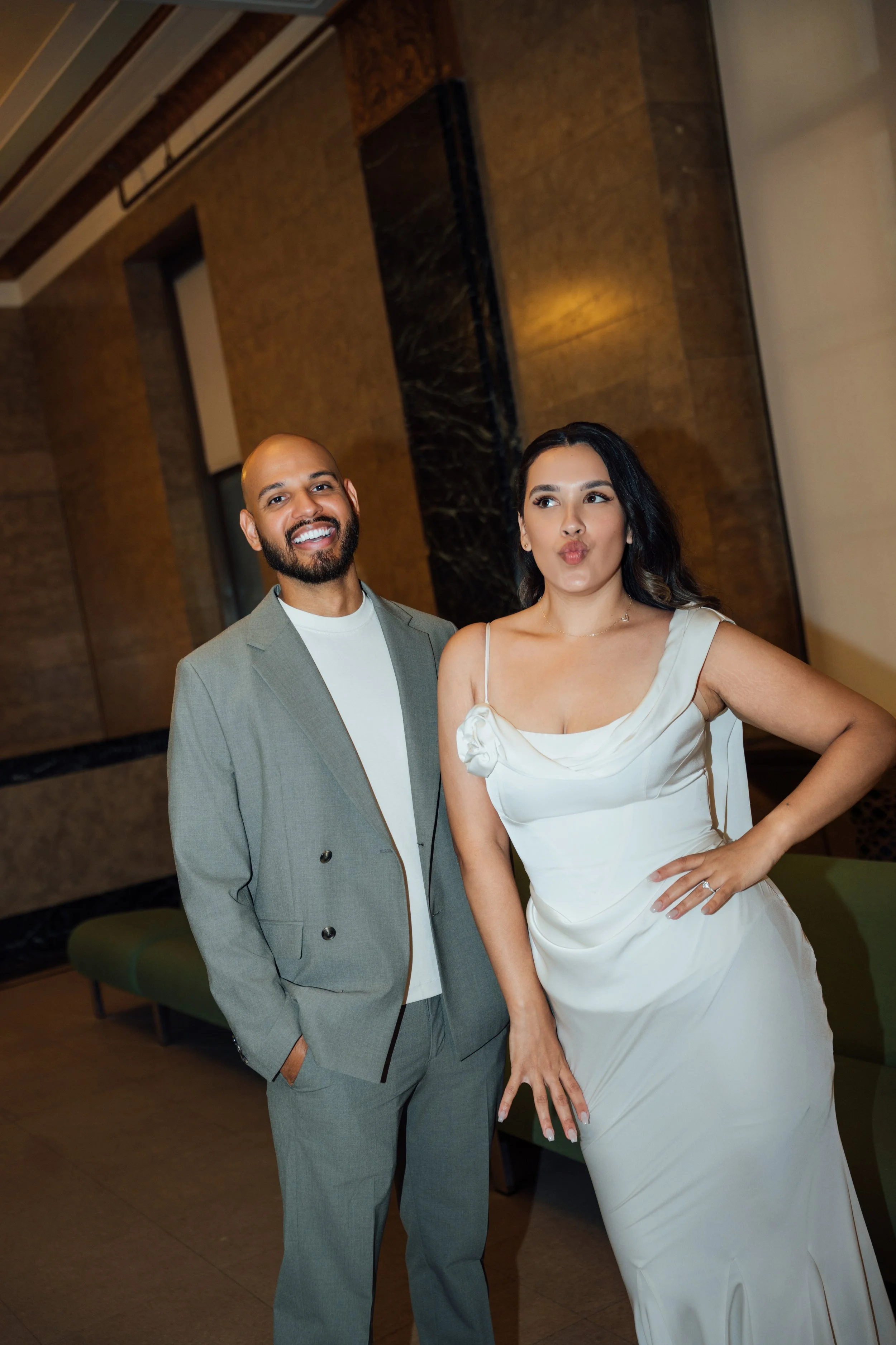 A man wearing a light gray suit with a white shirt standing next to a woman in a white dress. They are posing indoors with a warm-toned background, smiling and making playful faces.