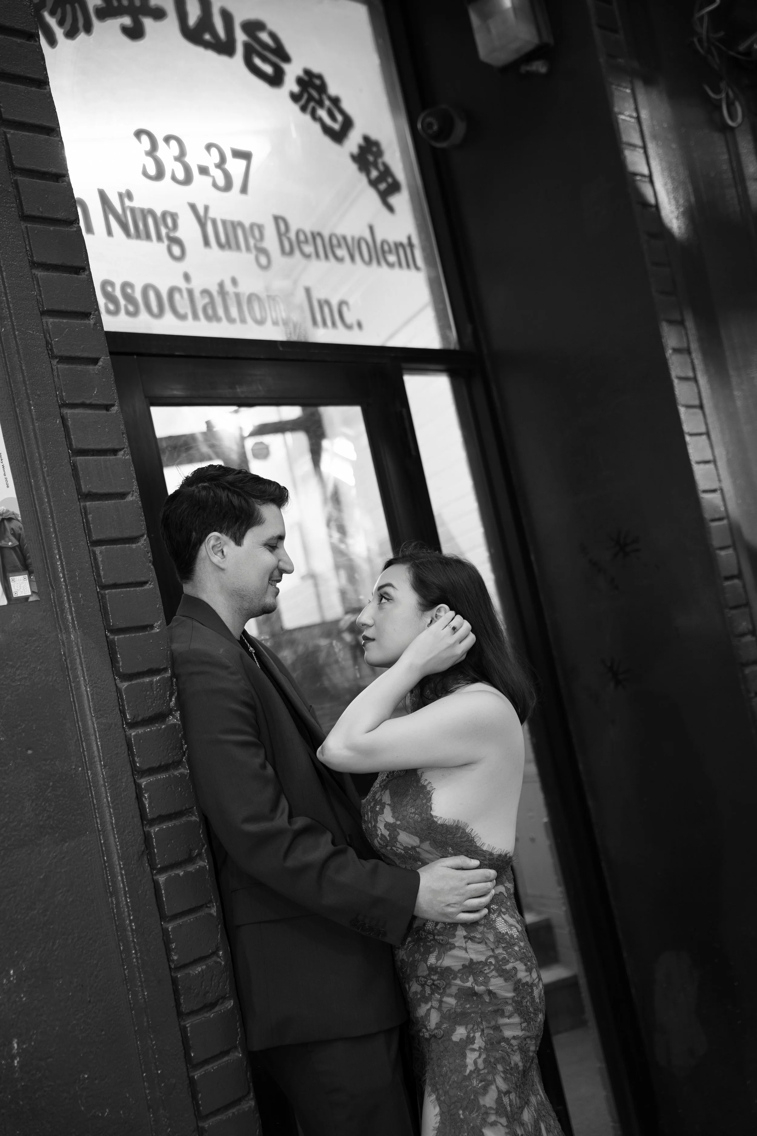 A man and woman are embracing and looking into each other's eyes outside a storefront with a sign displaying business hours and name in Asian characters.
