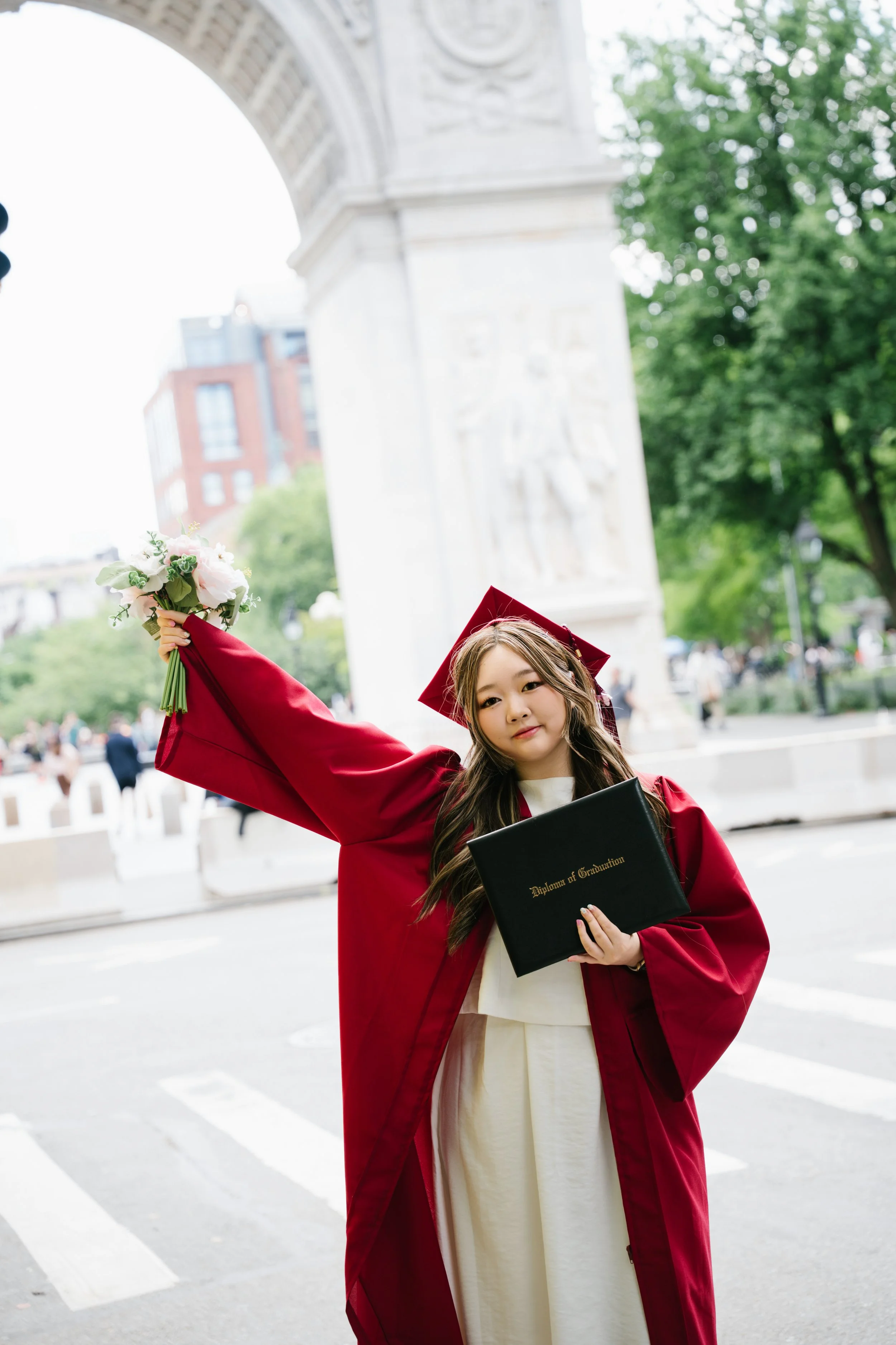 Young woman in a maroon graduation gown and cap holding a diploma and a bouquet of flowers outdoors near an arch and a sculpture, with trees and buildings in the background.