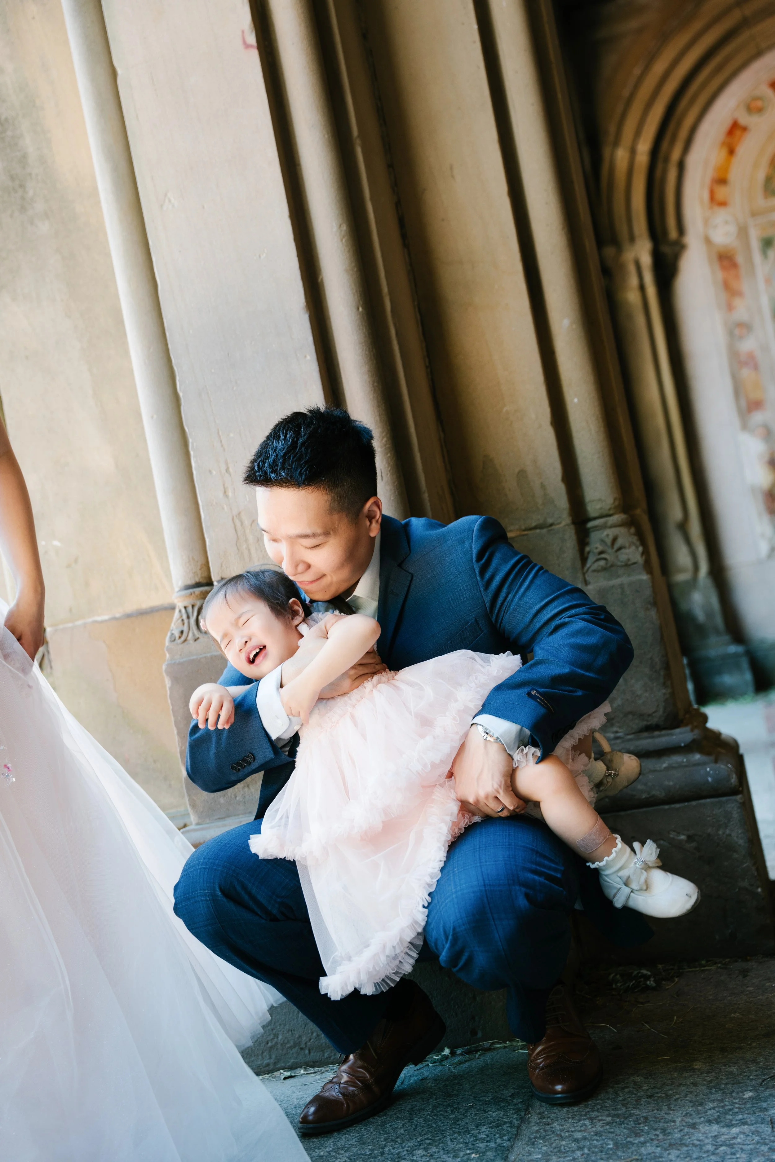 A man in a blue suit holding a laughing young girl in a pink dress, sitting on a stone step near a church interior with ornate arches and columns.