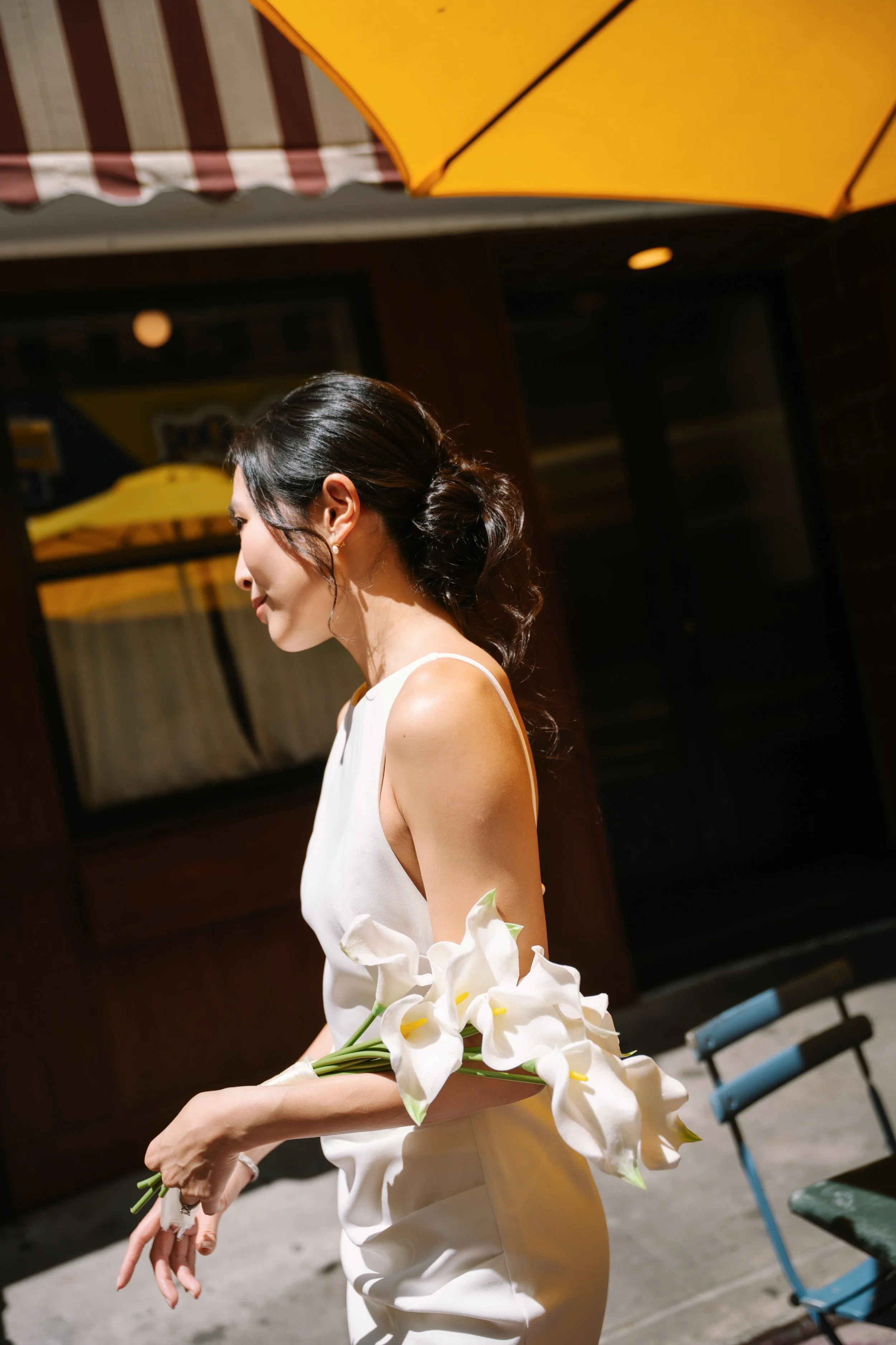 A woman with dark hair in an elegant updo, wearing a white dress, holding a bouquet of white calla lilies, standing outdoors near a yellow umbrella and a building at night.