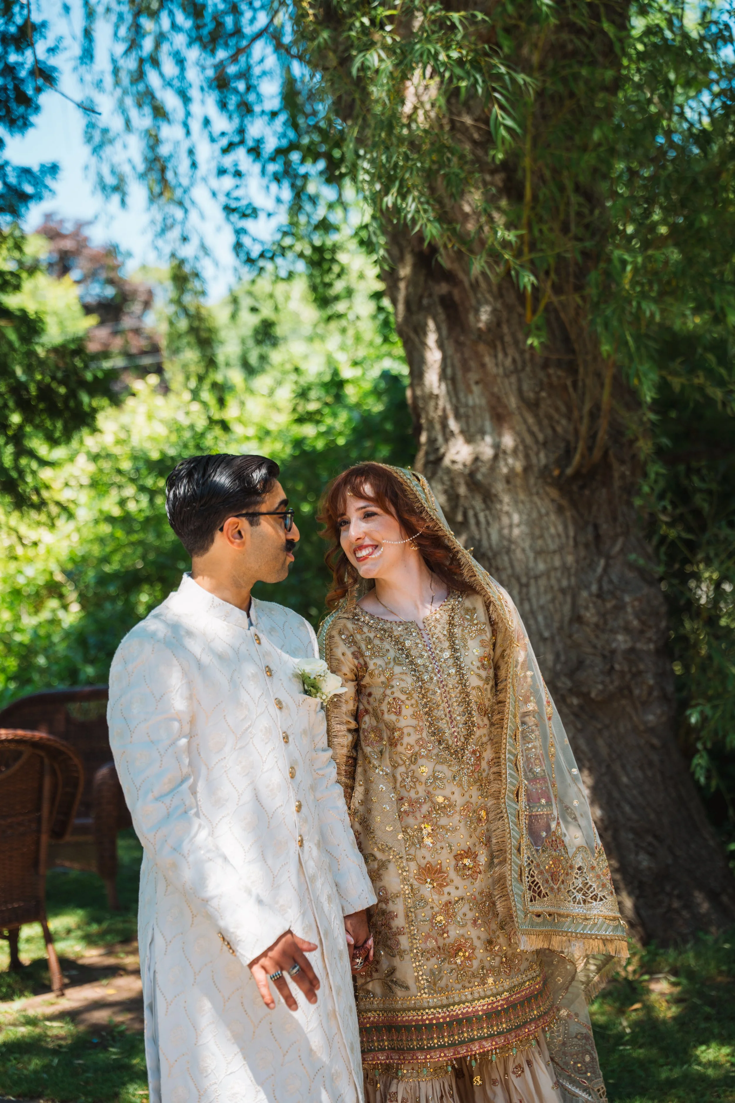 A couple dressed in traditional South Asian wedding attire standing outdoors in front of a large tree, smiling and looking at each other.