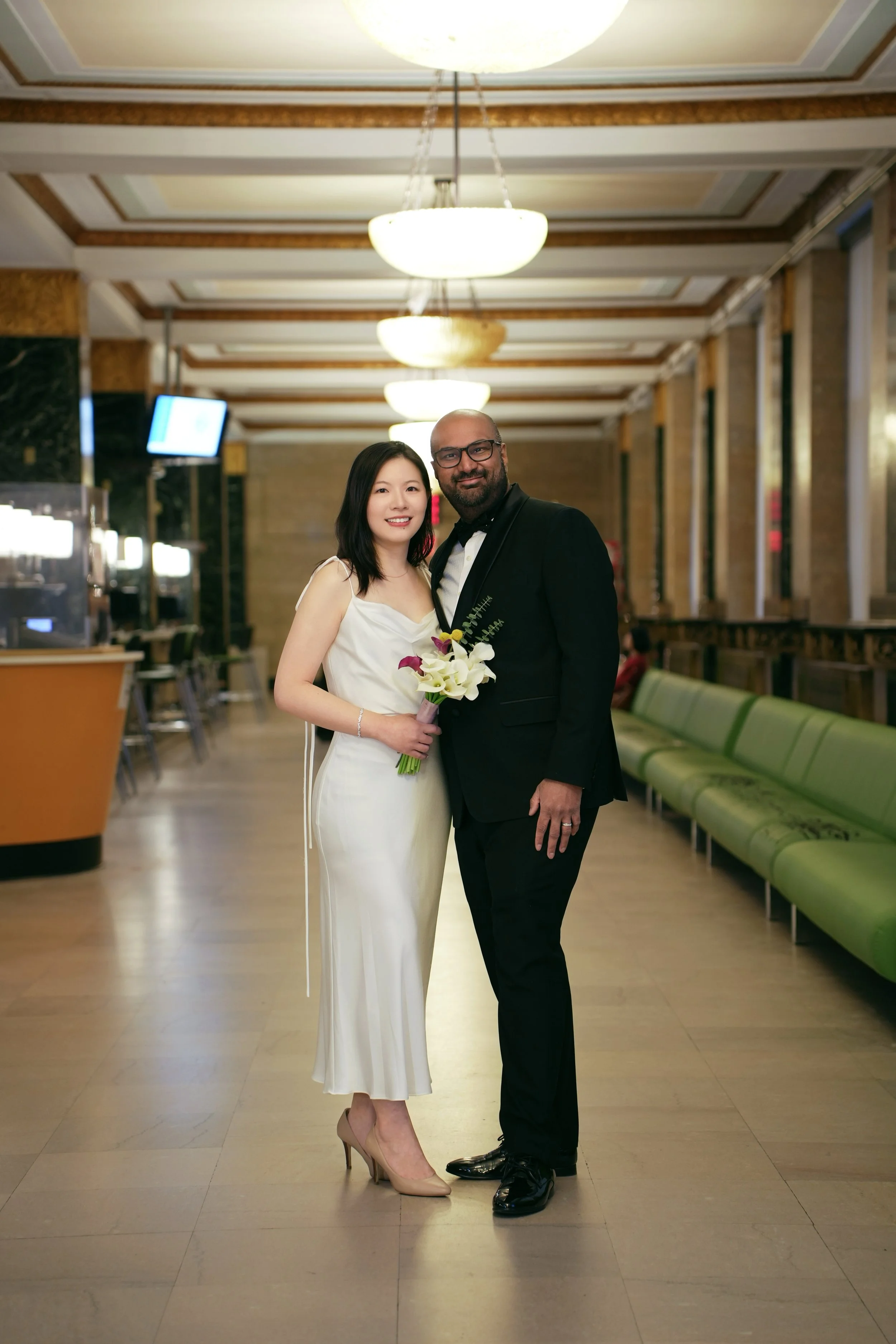 A smiling bride and groom stand inside a decorated hall. The bride wears a white dress and nude heels, holding a bouquet of flowers. The groom wears a black tuxedo with a white shirt and glasses.