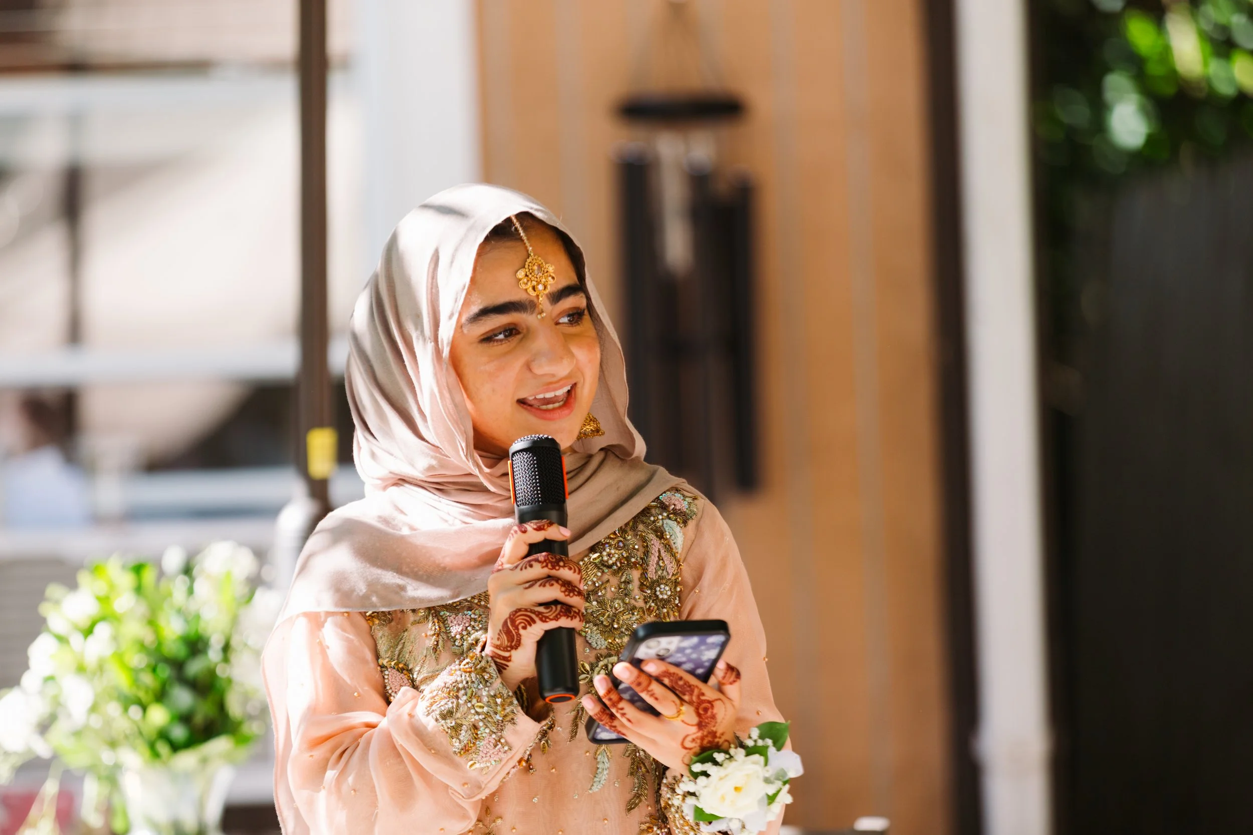 A young woman dressed in traditional South Asian attire, holding a microphone and a phone, with henna designs on her hands, standing outdoors with greenery in the background.
