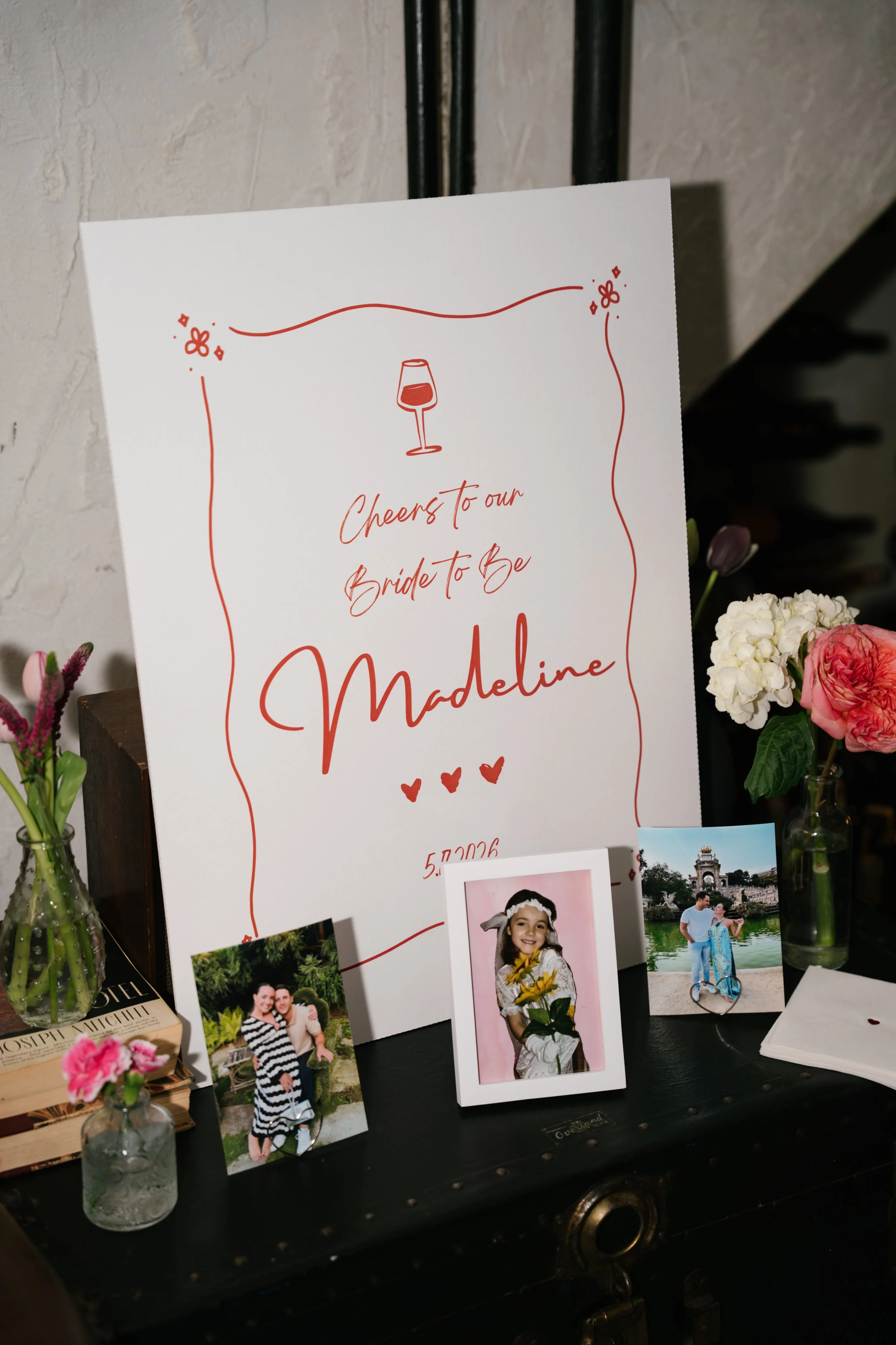 A display board with a sign that reads, 'Cheers to our Bride To Be Madeline,' along with a wine glass illustration. Surrounding the board are framed photos of smiling people, and small vases with flowers are also visible.