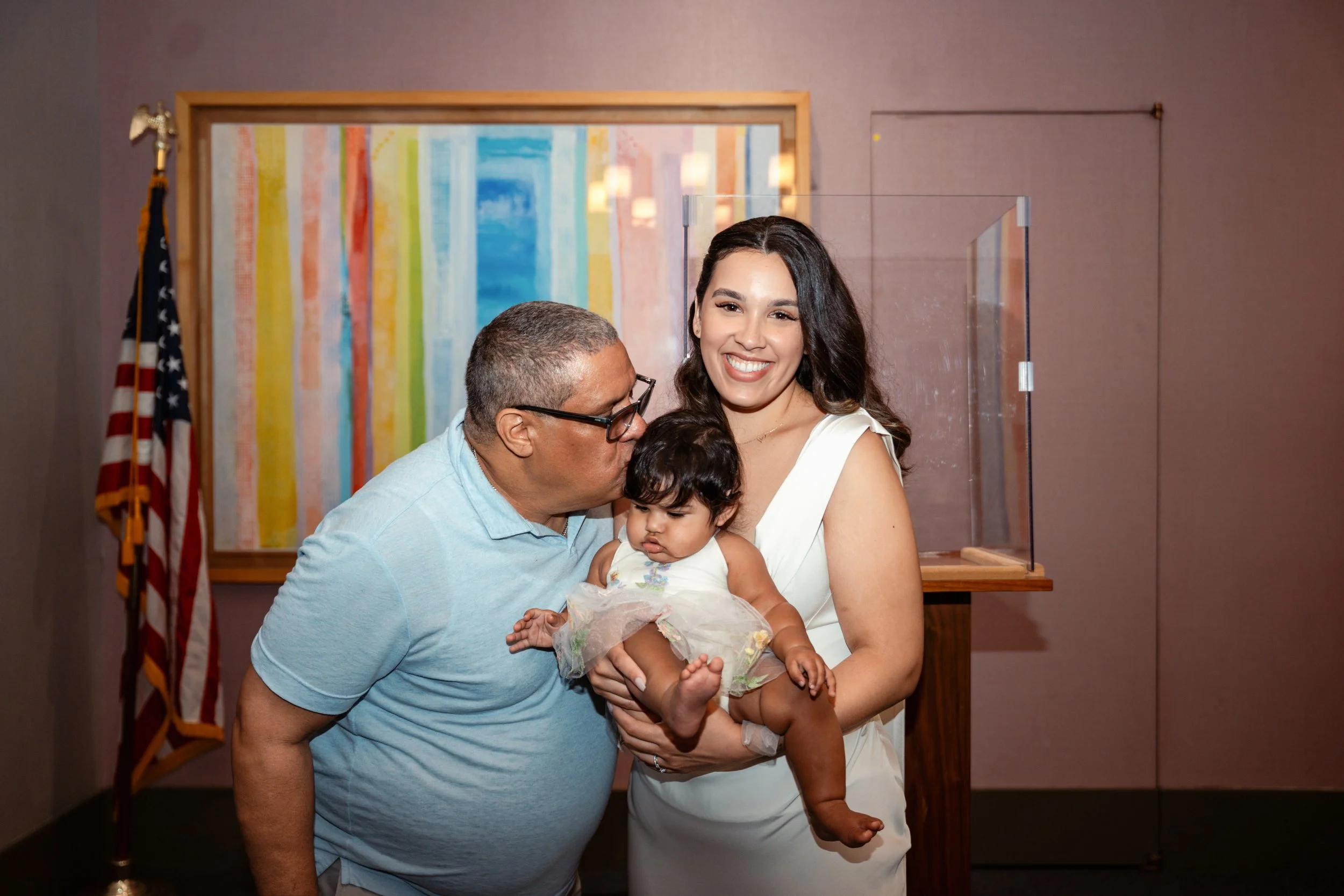 A woman holding a young girl, with an older man kissing the girl's forehead, at a ceremony or event.