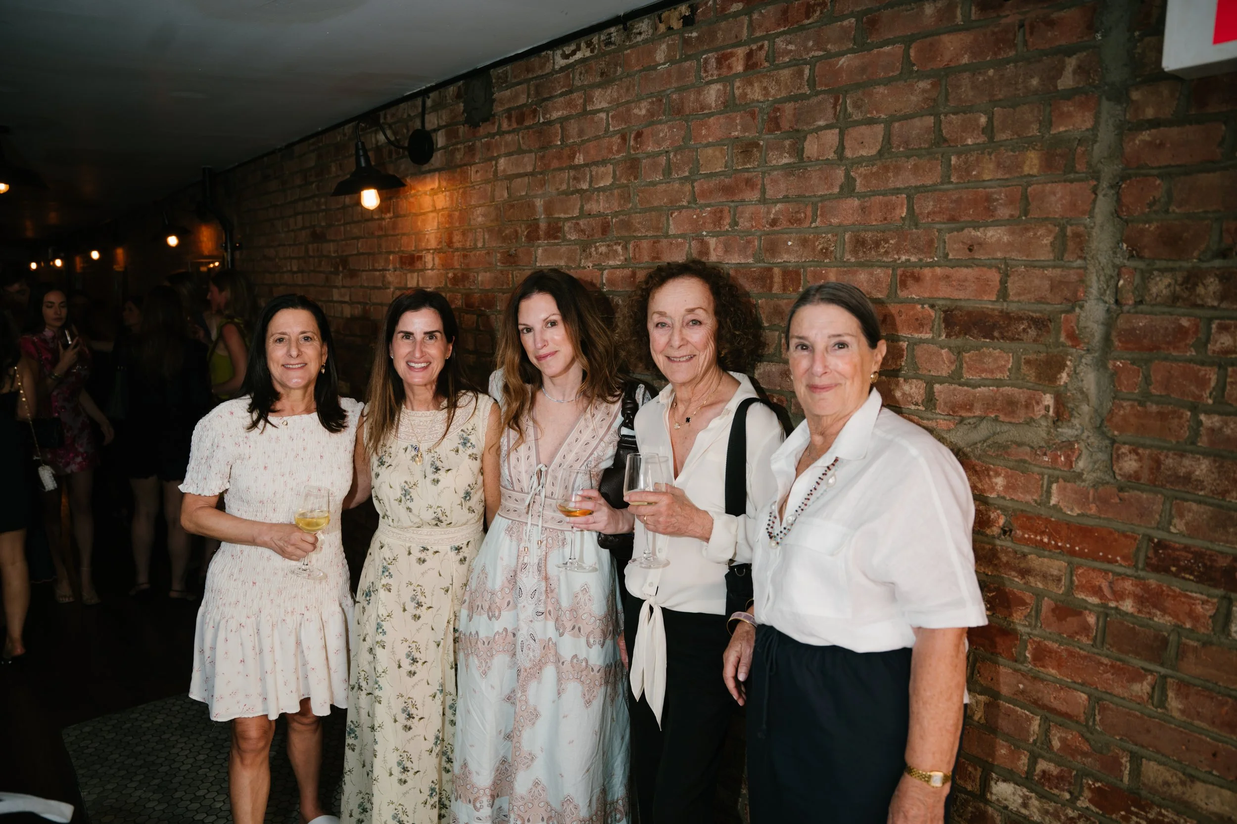 Five women standing in front of a brick wall, smiling, holding glasses with drinks at an indoor social event.