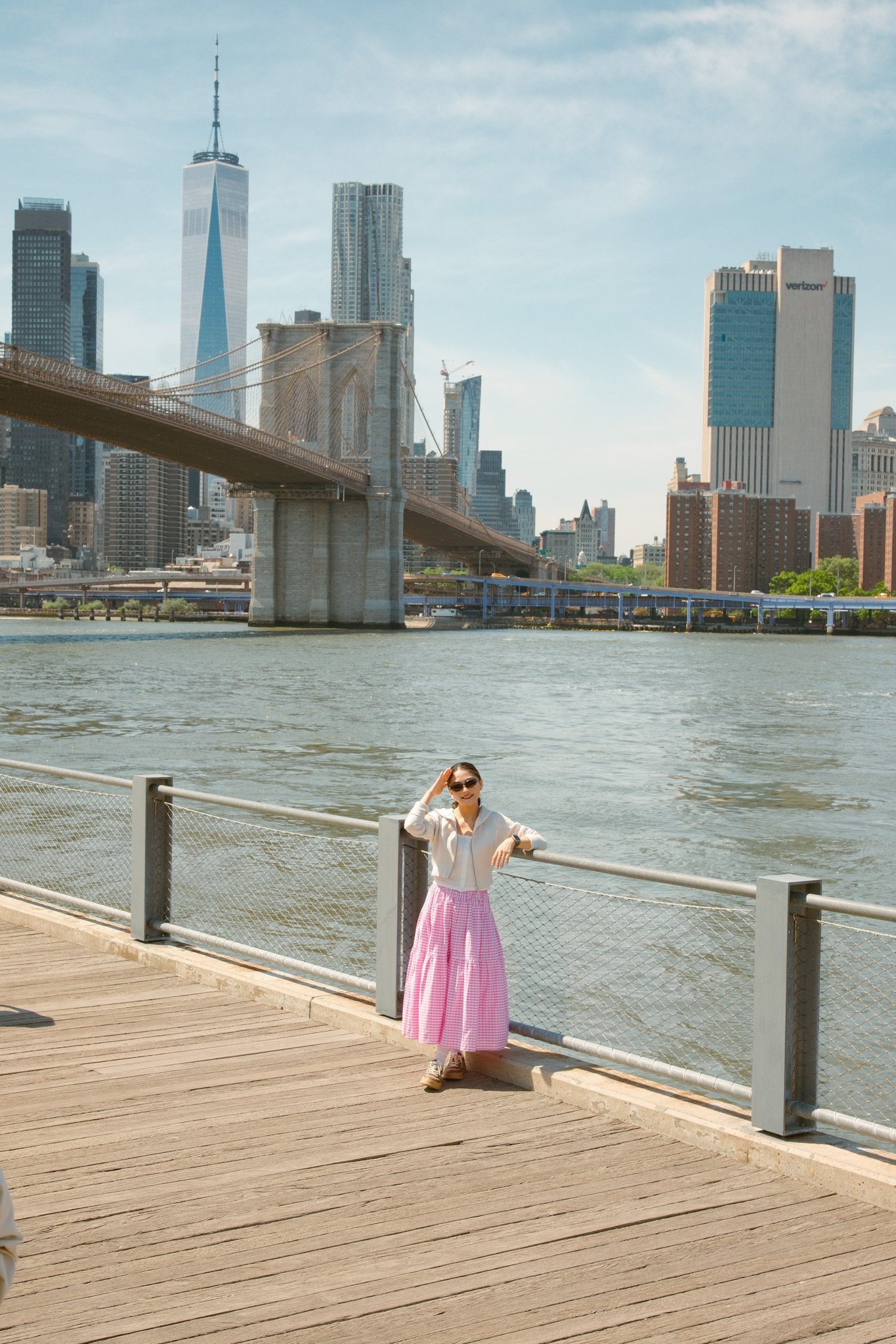 A woman standing on a wooden boardwalk by the water, smiling and wearing sunglasses, a white top, and a pink and white checkered skirt, with a city skyline including the Brooklyn Bridge and other tall buildings in the background.