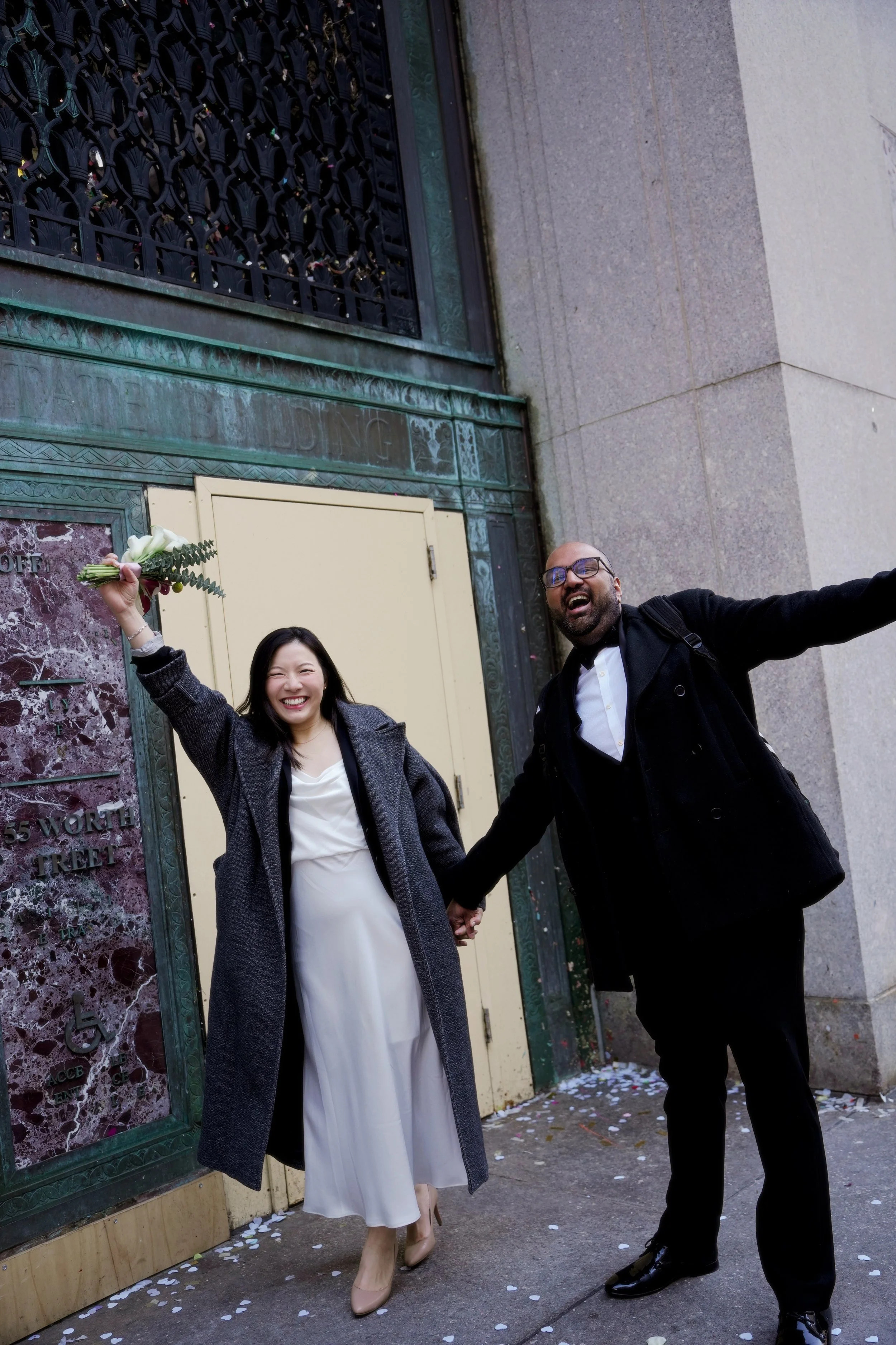 A joyful couple holding hands and smiling on a city sidewalk in front of a building with green metal ornate window guards and a beige door. The woman is wearing a white dress and beige heels, holding a small bouquet, and the man is dressed in a suit 