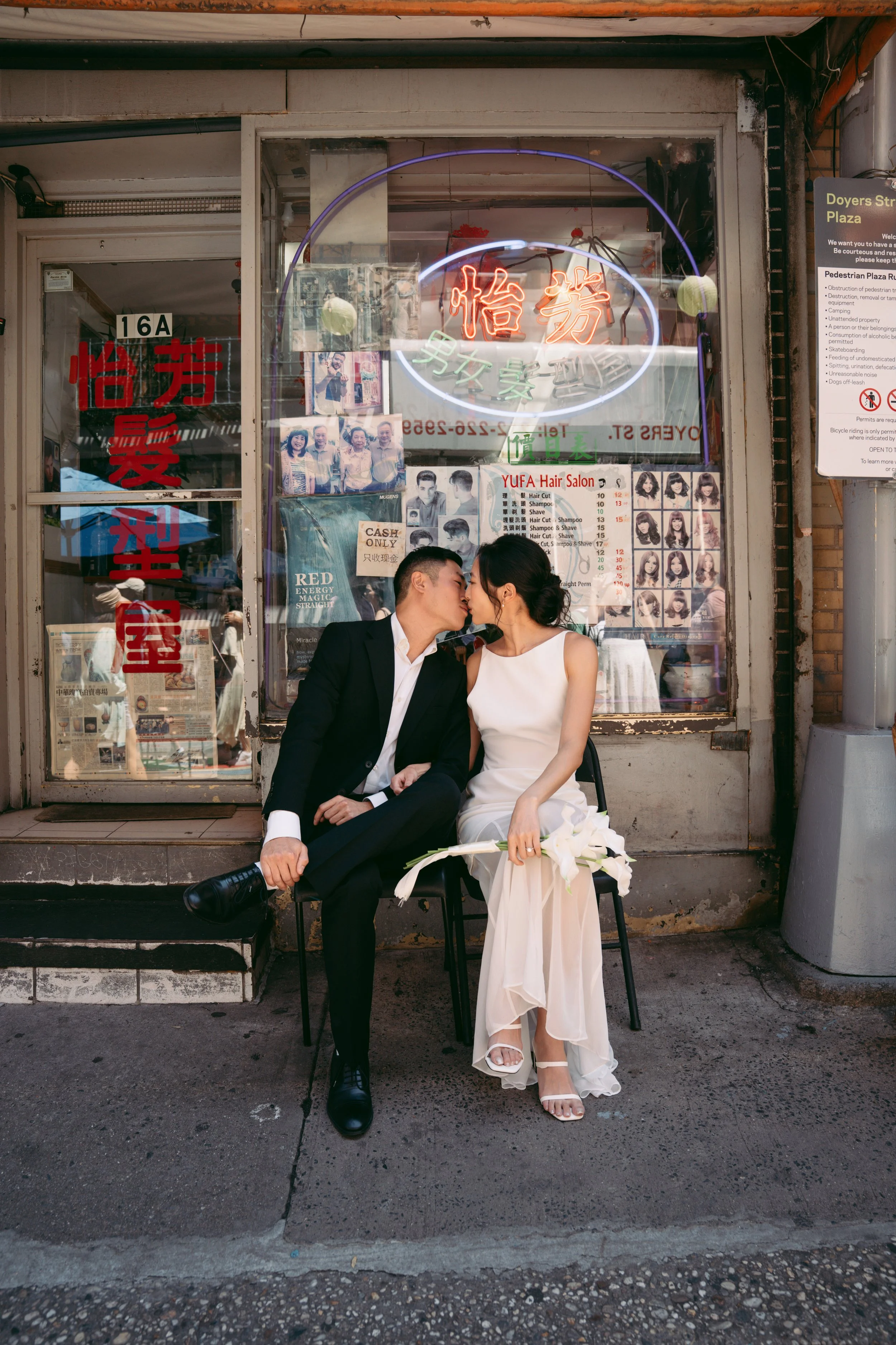 A couple in formal attire is sitting outside a storefront, about to share a kiss. The man is in a black suit and white shirt, while the woman is in a white dress and heels, holding a bouquet of white flowers. The storefront behind them has neon signs